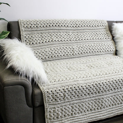 May include: A light gray crocheted throw blanket draped over a gray couch with a white faux fur pillow. The blanket has a textured stitch pattern.