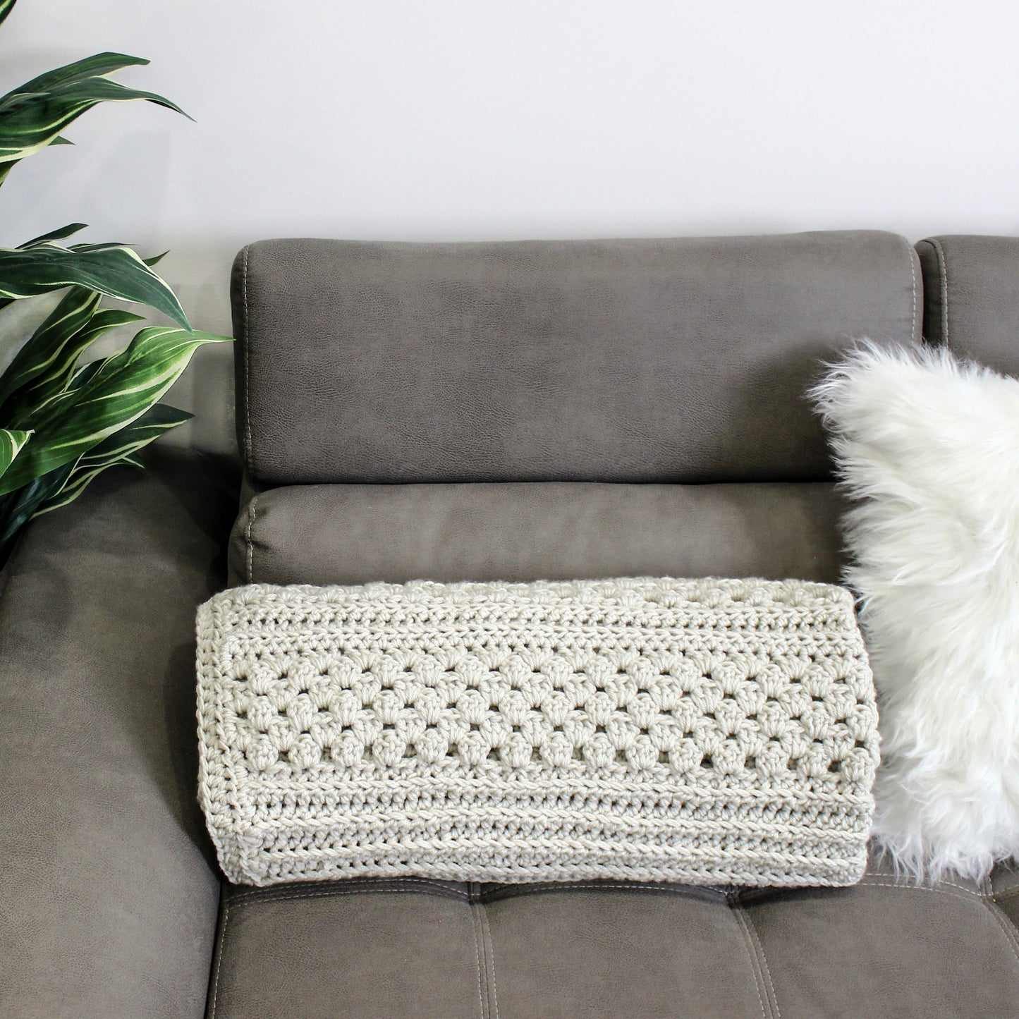 May include: A light gray crocheted throw blanket is draped over a gray couch with a white faux fur pillow.