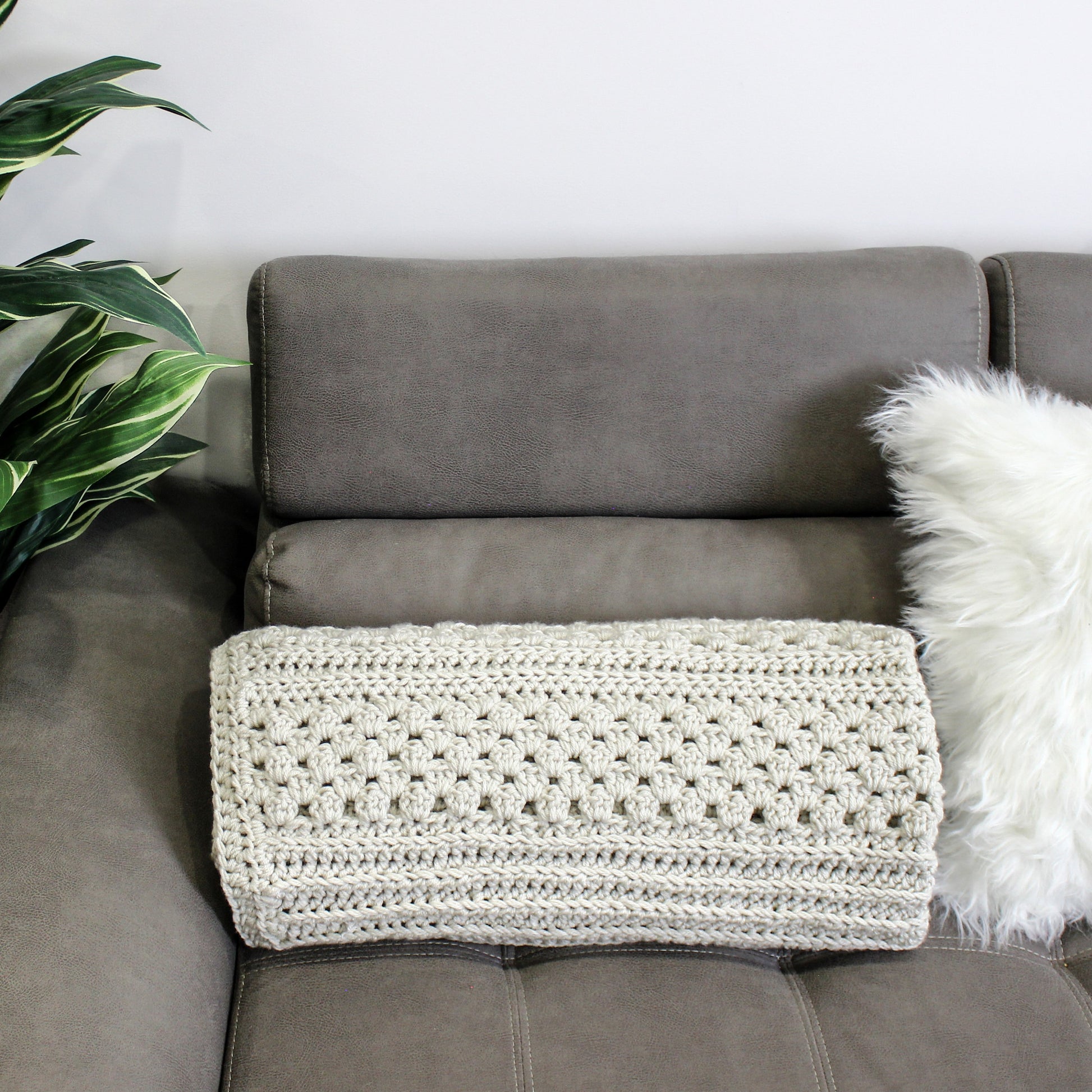 May include: A light gray crocheted throw blanket is draped over a gray couch with a white faux fur pillow.