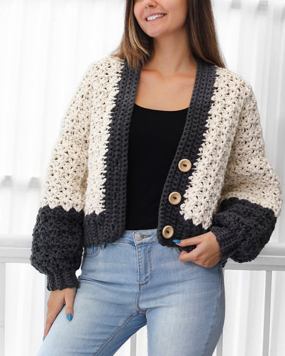 May include: A cropped cardigan with a white and gray color block design. The cardigan has a button closure and long sleeves. The sleeves are puffed at the shoulders and have a textured knit.