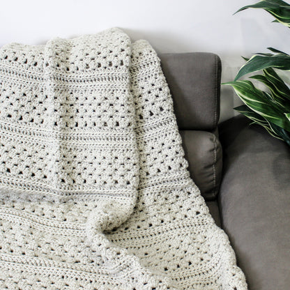 May include: A white crocheted throw blanket draped over a gray couch. The blanket has a textured, open-stitch pattern.