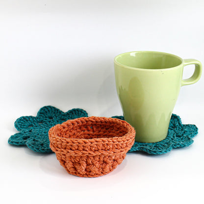 May include: A crocheted teal coaster with a crocheted orange bowl on top. A light green ceramic mug sits on the coaster.