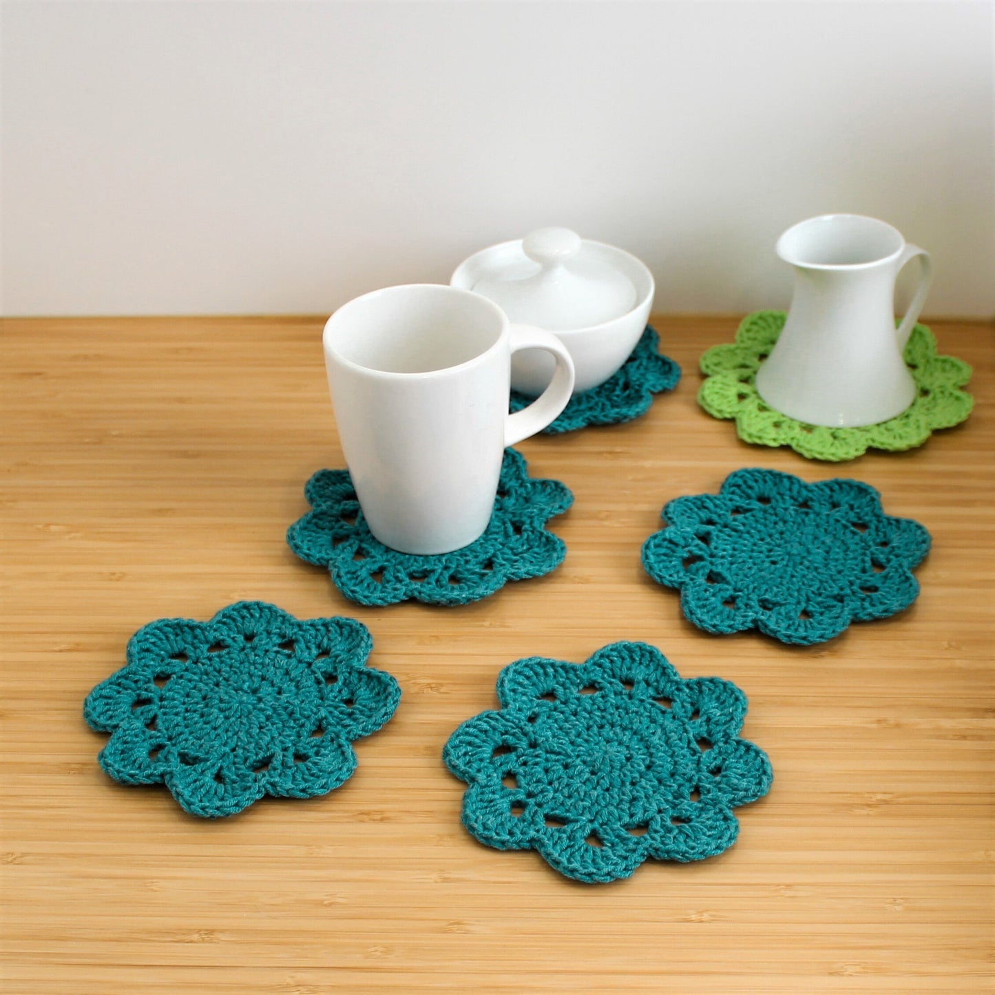 May include: Five crocheted coasters in teal and green. The coasters are in the shape of a flower with multiple layers of crocheted stitches. Two of the coasters have white ceramic mugs and a sugar bowl on top.