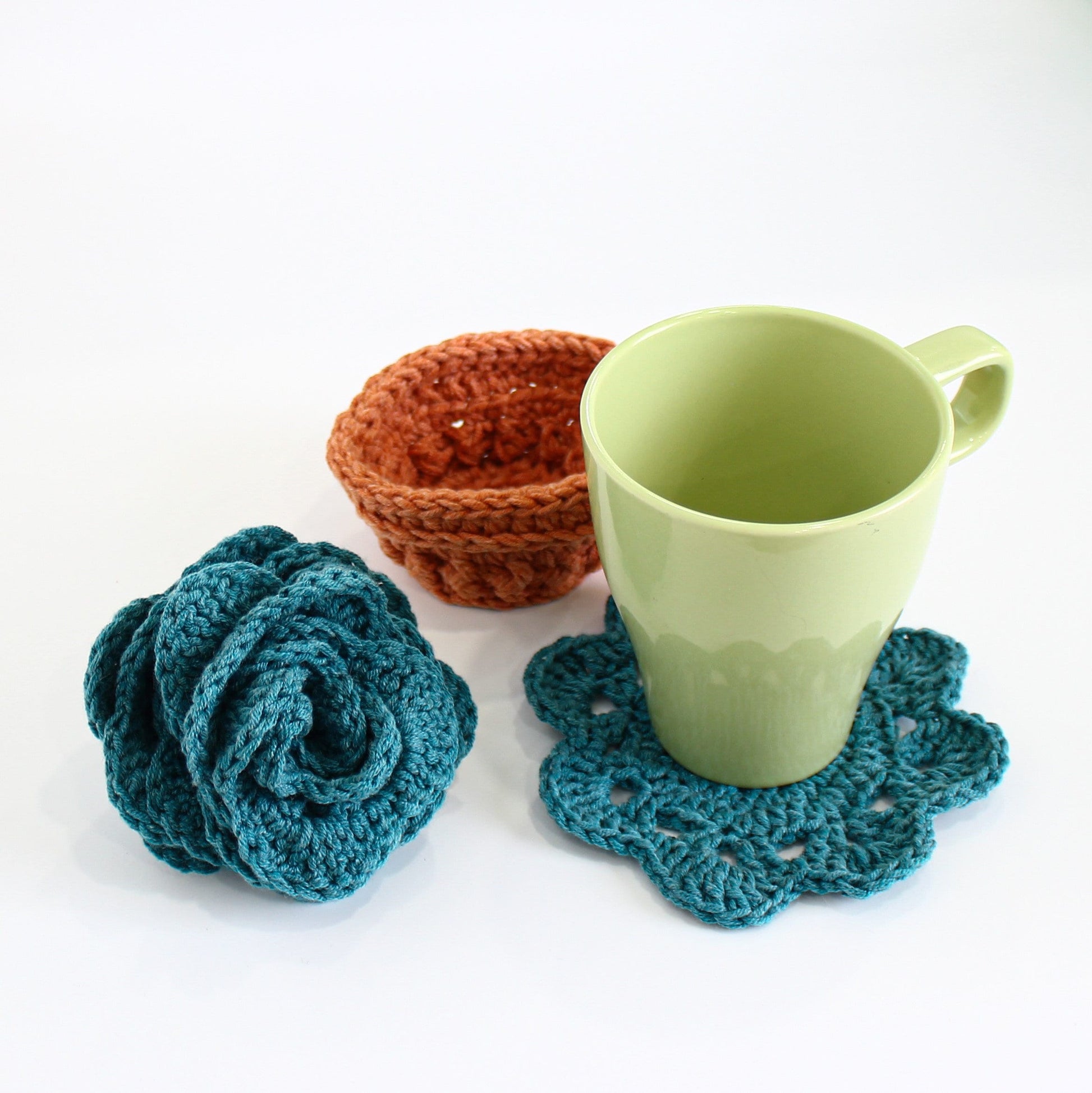 May include: A set of three crocheted items: a teal blue flower, a teal blue coaster, and a brown crocheted bowl. The coaster is under a light green ceramic mug.