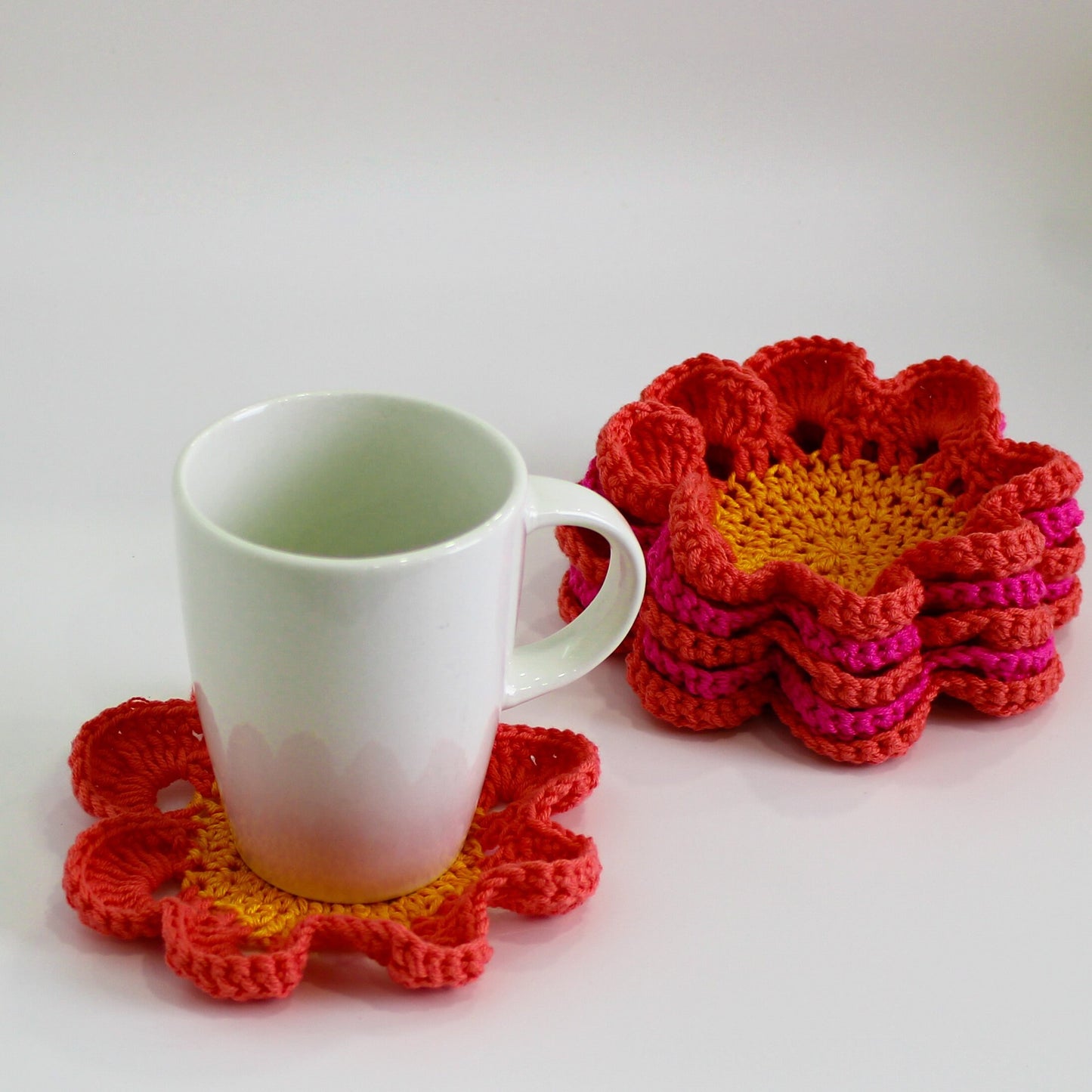 May include: A white ceramic mug sits on a crocheted flower-shaped coaster. The coaster is red and yellow and has a layered design. There are three more crocheted coasters in the background, stacked on top of each other.