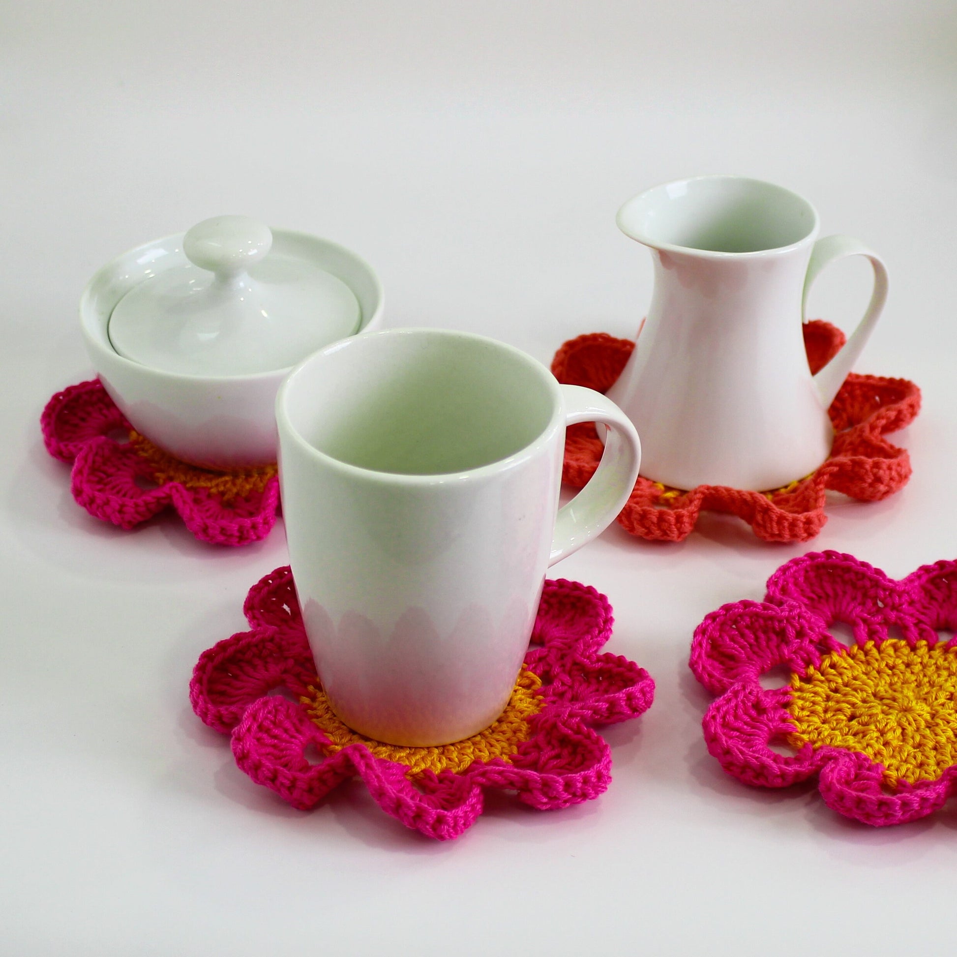 May include: Three white ceramic cups and a sugar bowl with lids, sitting on crocheted flower-shaped coasters in pink, orange, and yellow.