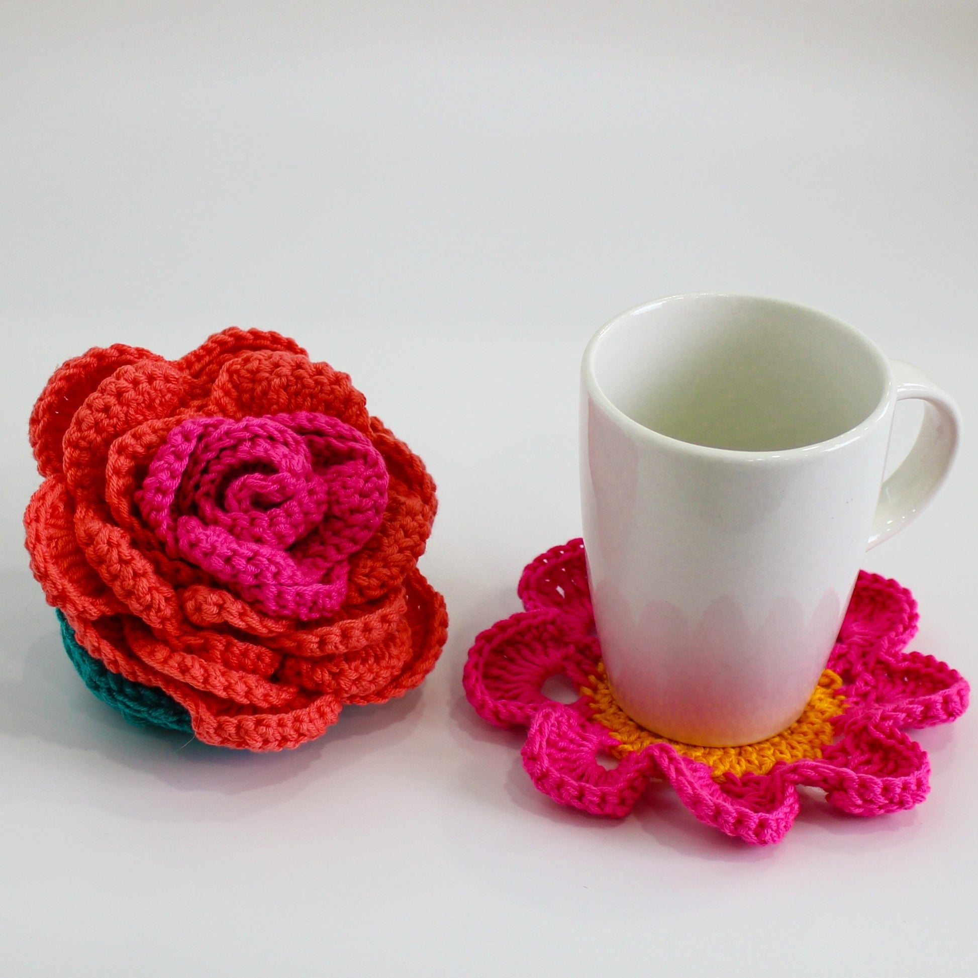 May include: A white ceramic mug with a crocheted coaster featuring pink and yellow flowers. A crocheted flower in shades of pink, red, and green is also pictured.