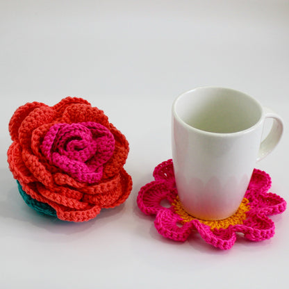 May include: A white ceramic mug with a crocheted coaster featuring pink and yellow flowers. A crocheted flower in shades of pink, red, and green is also pictured.
