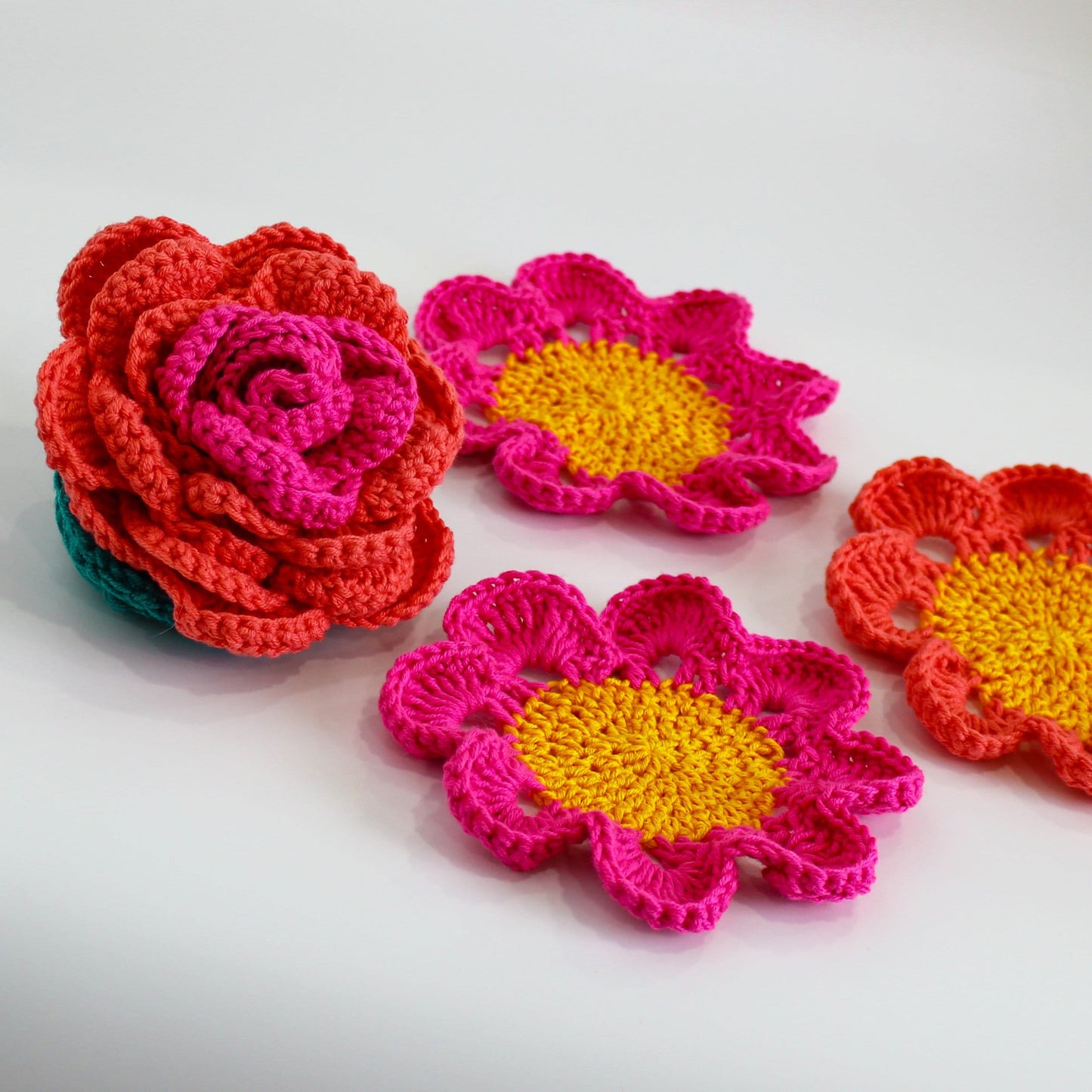 May include: Three crocheted flower coasters in pink, orange, and yellow. The coasters are in the shape of flowers with a center circle.