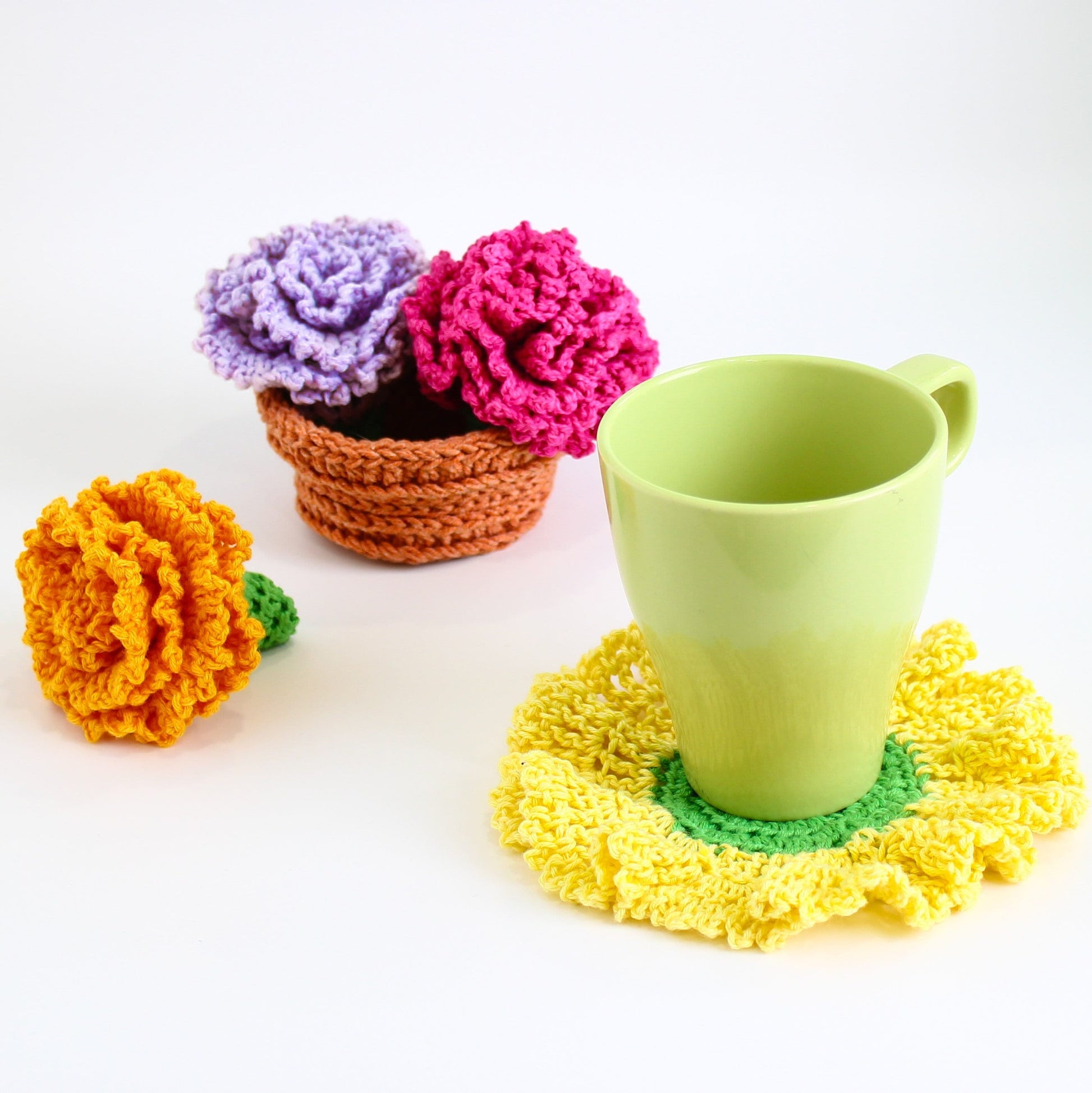 May include: A crocheted flower coaster in yellow, green, and white with a green mug on top. There are three crocheted flowers in a brown pot in the background.