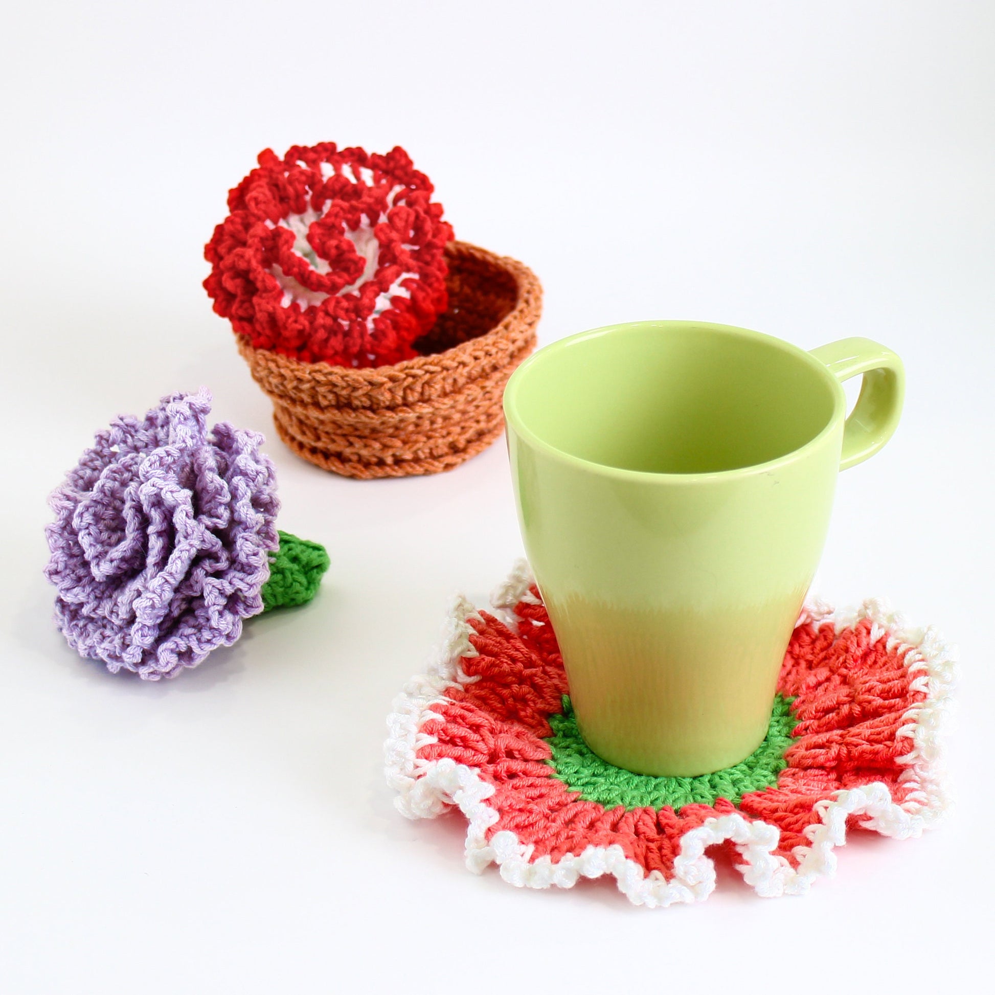 May include: A crocheted coaster in the shape of a flower with red, green, and white accents. A green ceramic mug sits on top of the coaster. Two crocheted carnations, one red and one purple, are in the background.