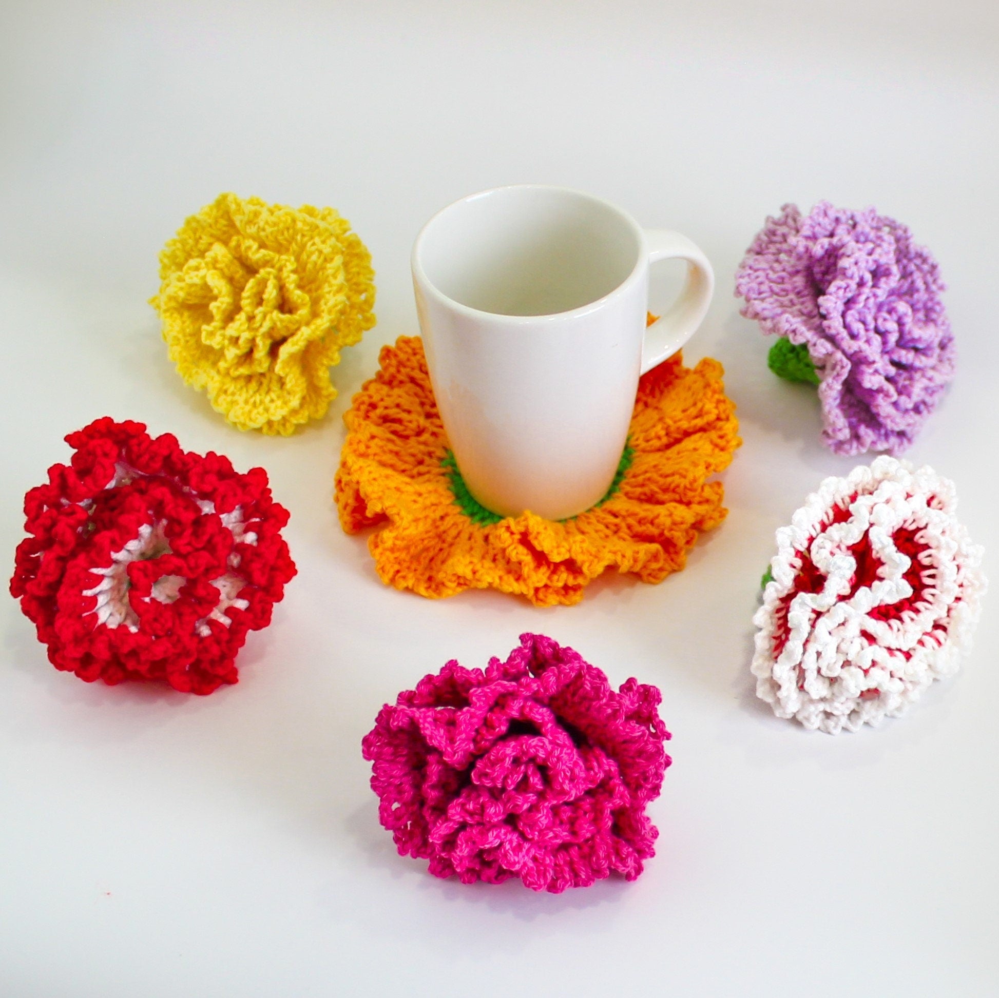 May include: Five crocheted carnation flower coasters in red, yellow, pink, white, and purple, with a white ceramic mug sitting on the orange coaster.