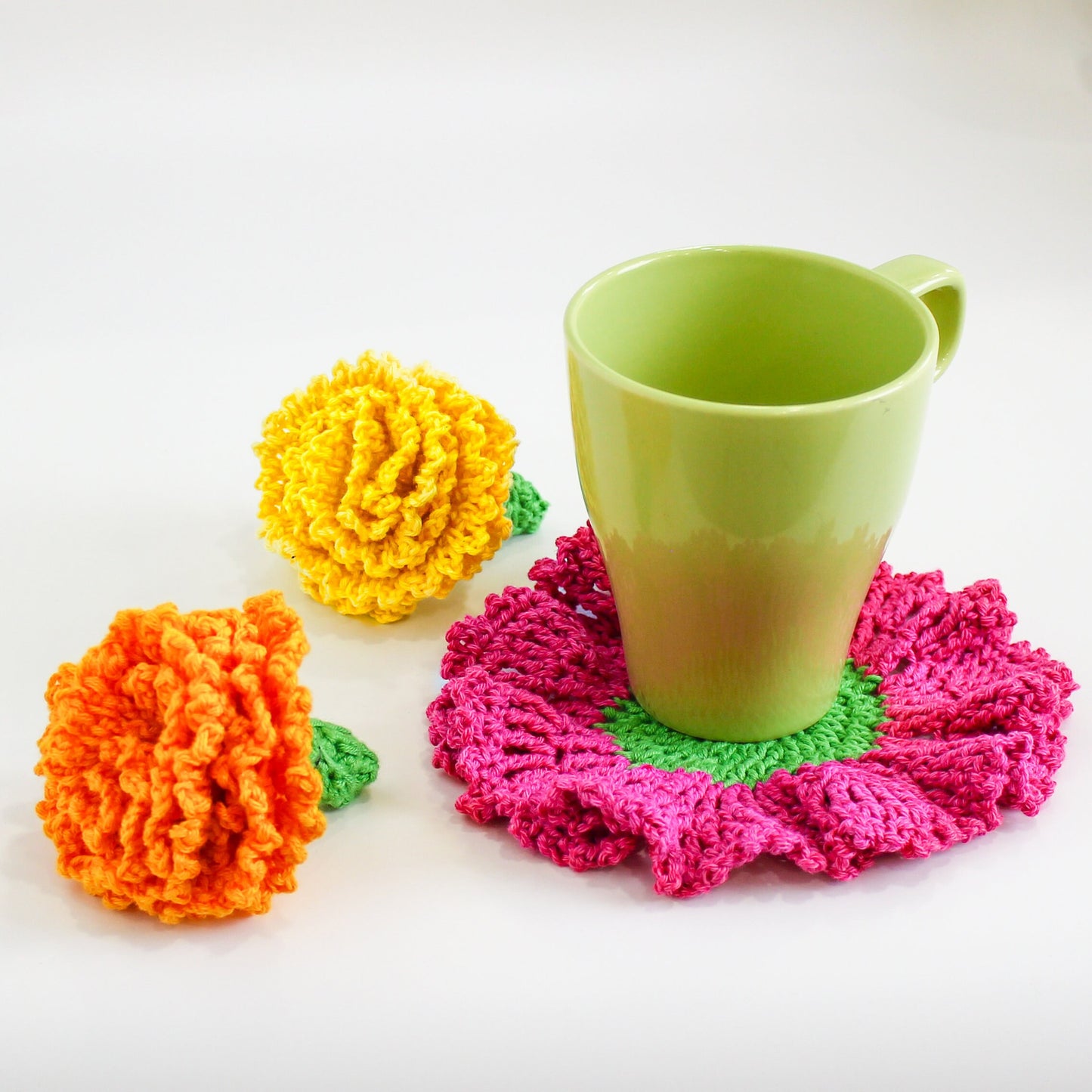 May include: A green ceramic mug sits on a crocheted coaster with a pink and green flower design. Two crocheted flowers, one yellow and one orange, are on the white surface.