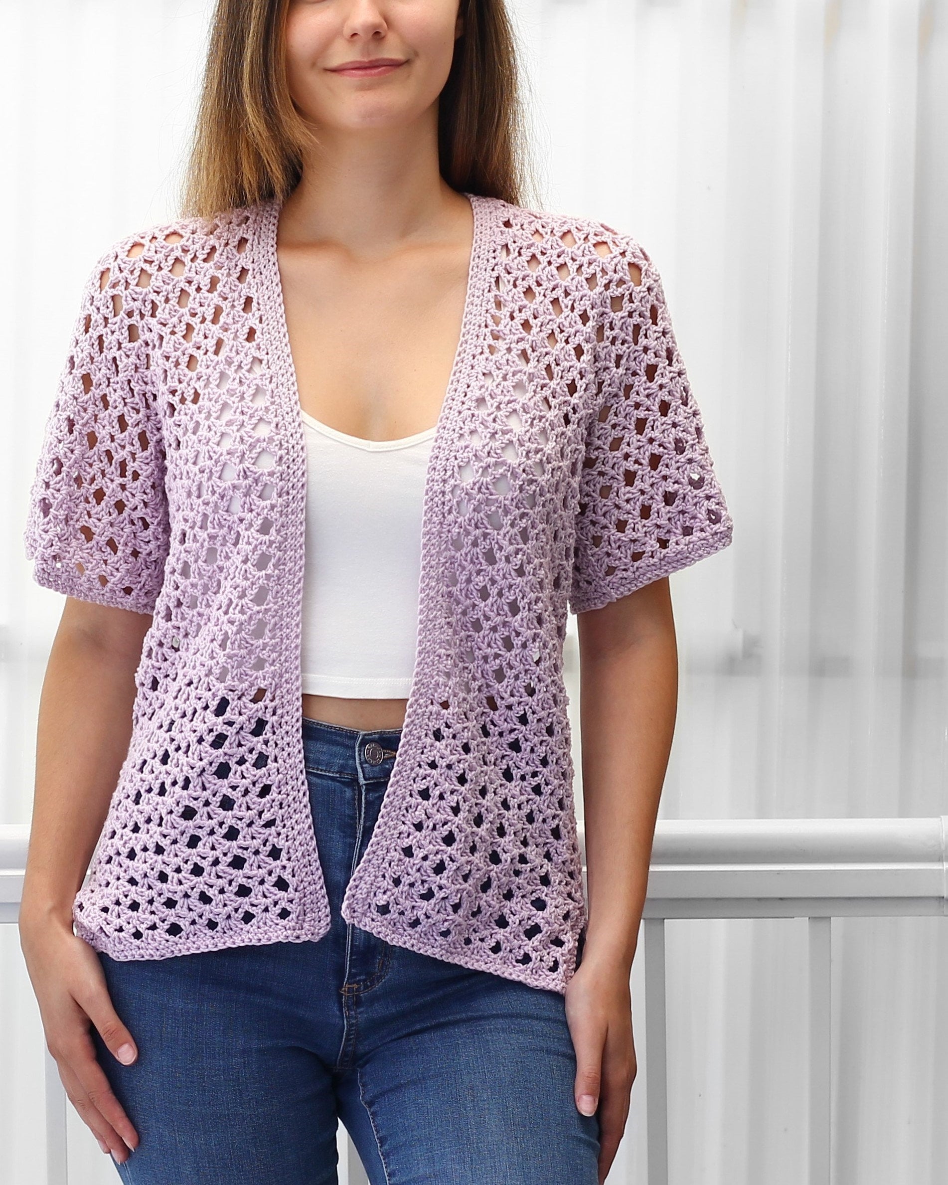May include: A light purple crochet cardigan with a V-neck and short sleeves. The cardigan is made with a delicate open stitch pattern.
