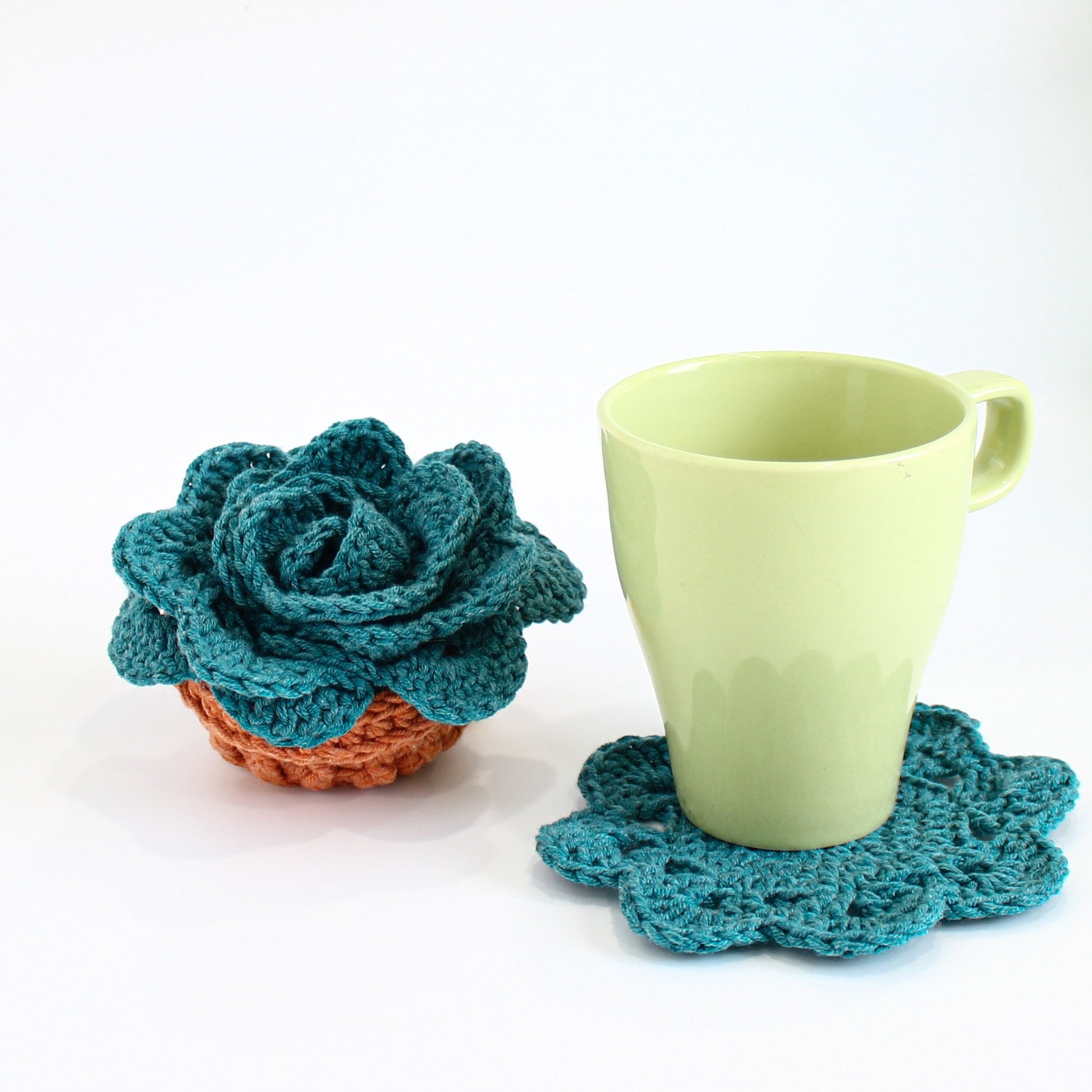 May include: A teal crocheted succulent plant in a terracotta pot next to a light green mug on a teal crocheted coaster.