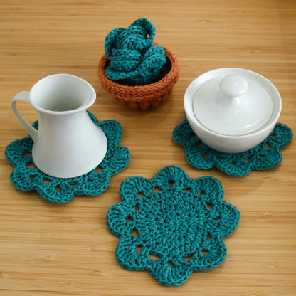 May include: Three teal crocheted coasters with a white ceramic pitcher and a white ceramic sugar bowl with a lid on a wooden surface.
