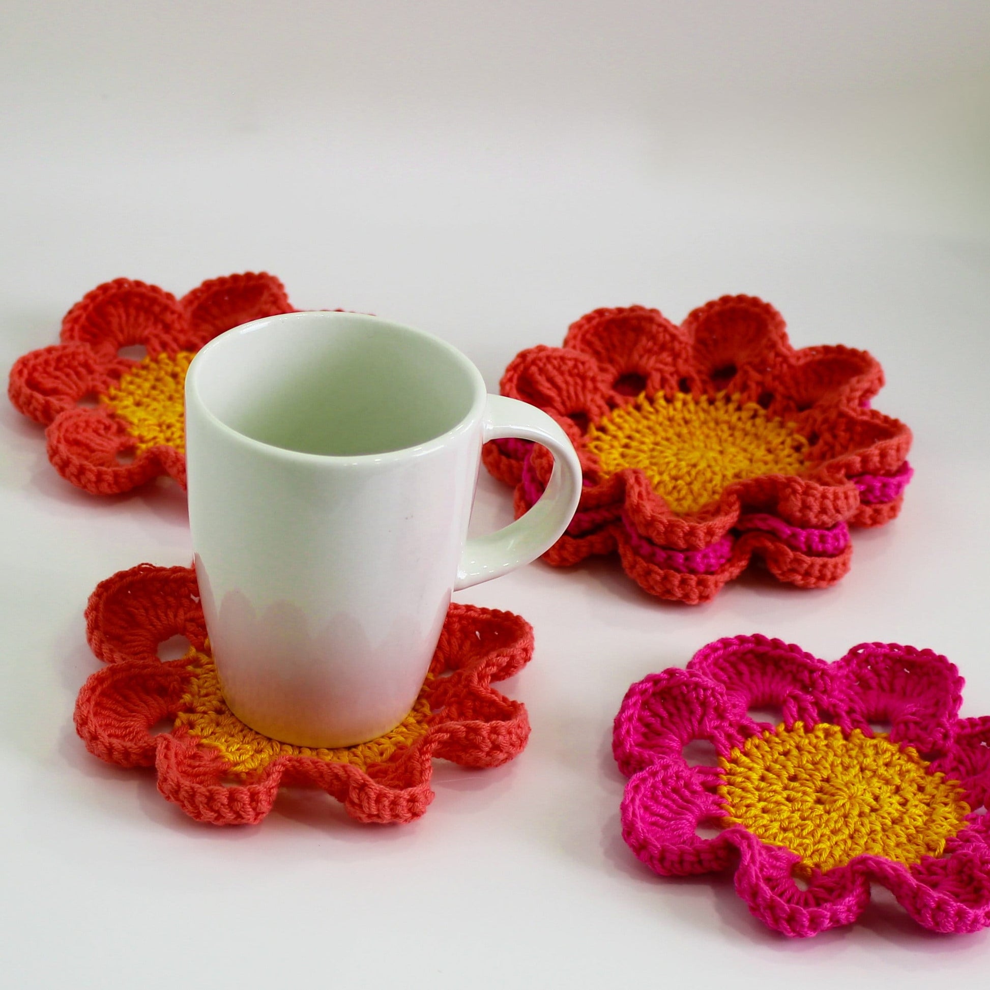 May include: Four crocheted flower-shaped coasters in pink and yellow. A white ceramic mug sits on one of the coasters.