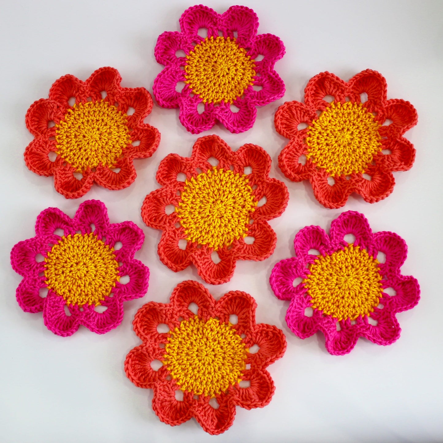 May include: Six crocheted flower coasters in shades of pink and yellow. The coasters are made with a textured yarn and have a raised center.