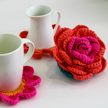 May include: Two white ceramic mugs with crocheted coasters. One coaster is a bright pink and yellow flower, the other is a large, multi-colored crocheted flower.