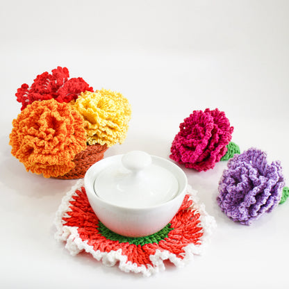 May include: A set of crocheted carnation flowers in various colors, including orange, yellow, pink, and purple. The flowers are arranged around a white ceramic sugar bowl with a lid. The flowers are sitting on a crocheted coaster.