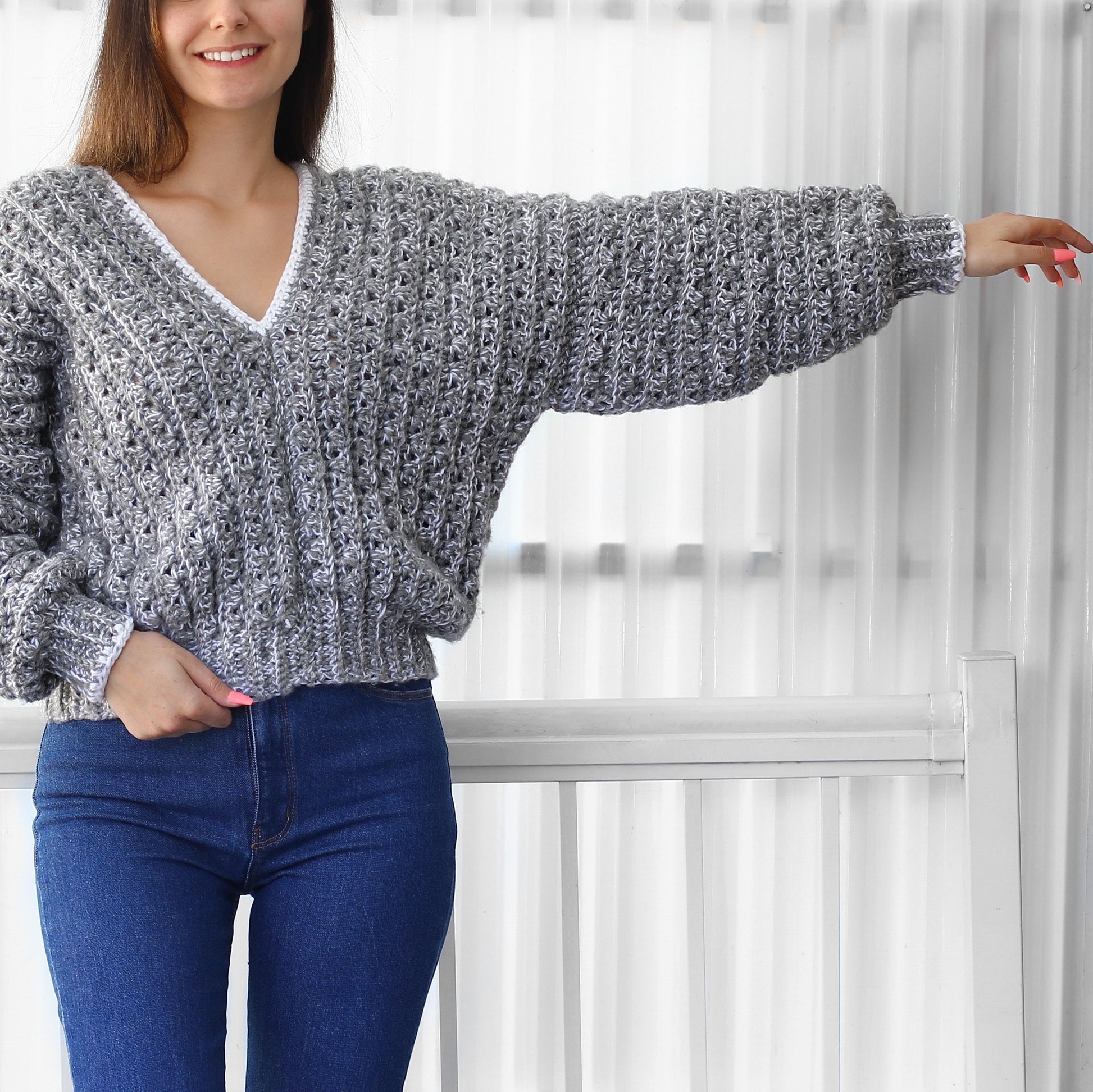May include: A gray crocheted sweater with a white trim around the neckline and sleeves. The sweater has a loose fit and a V-neck.