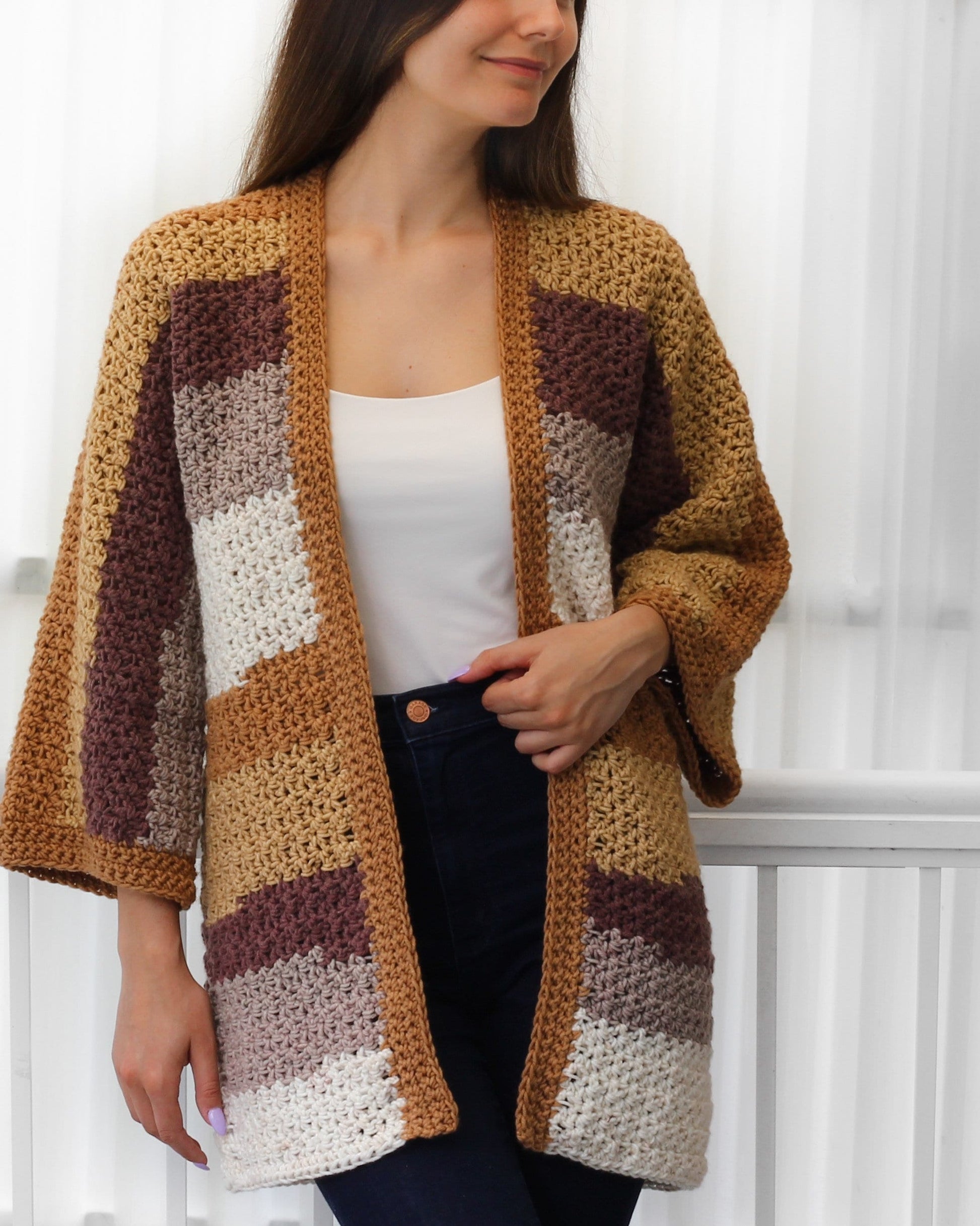 May include: A handmade, open-front crochet cardigan in a patchwork design. The color scheme includes shades of tan, brown, and cream. The sleeves are three-quarter length. The cardigan is a cozy, stylish garment.