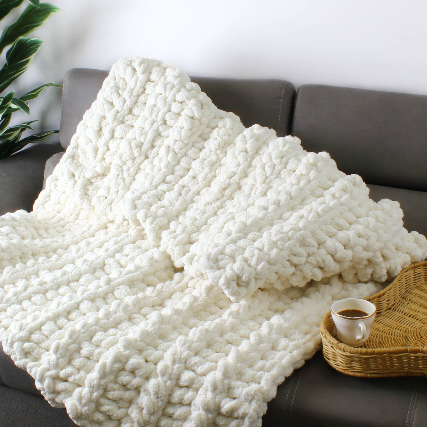 May include: A white chunky knit throw blanket draped over a gray couch with a cup of coffee and a wicker tray on the couch.