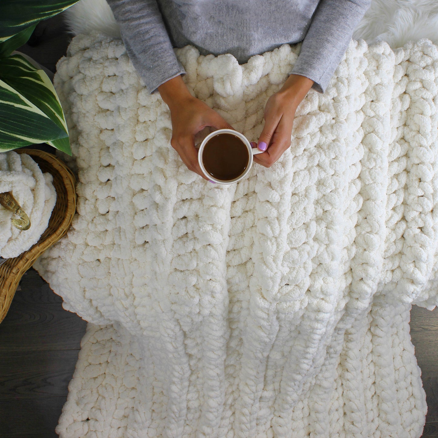 May include: A white chunky knit blanket with a person holding a white mug of coffee. The blanket is made of thick yarn and has a soft, fluffy texture.