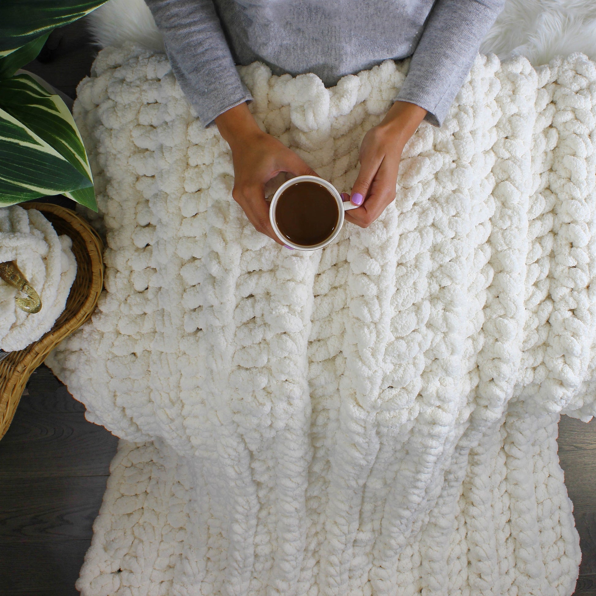 May include: A white chunky knit blanket with a person holding a white mug of coffee. The blanket is made of thick yarn and has a soft, fluffy texture.