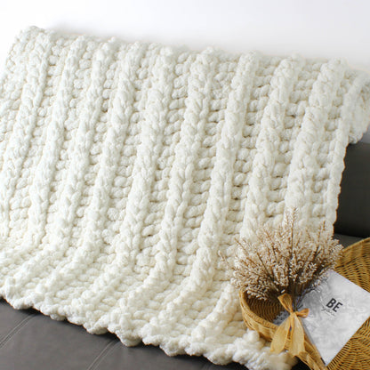 May include: A white chunky knit throw blanket with a soft, fluffy texture. The blanket is draped over a couch and has a natural, relaxed look.