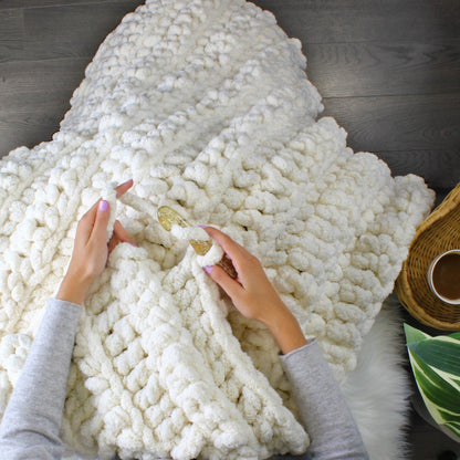 May include: A person is crocheting a white chunky knit blanket with large pom poms. The blanket is soft and fluffy, and the person is using a large crochet hook.