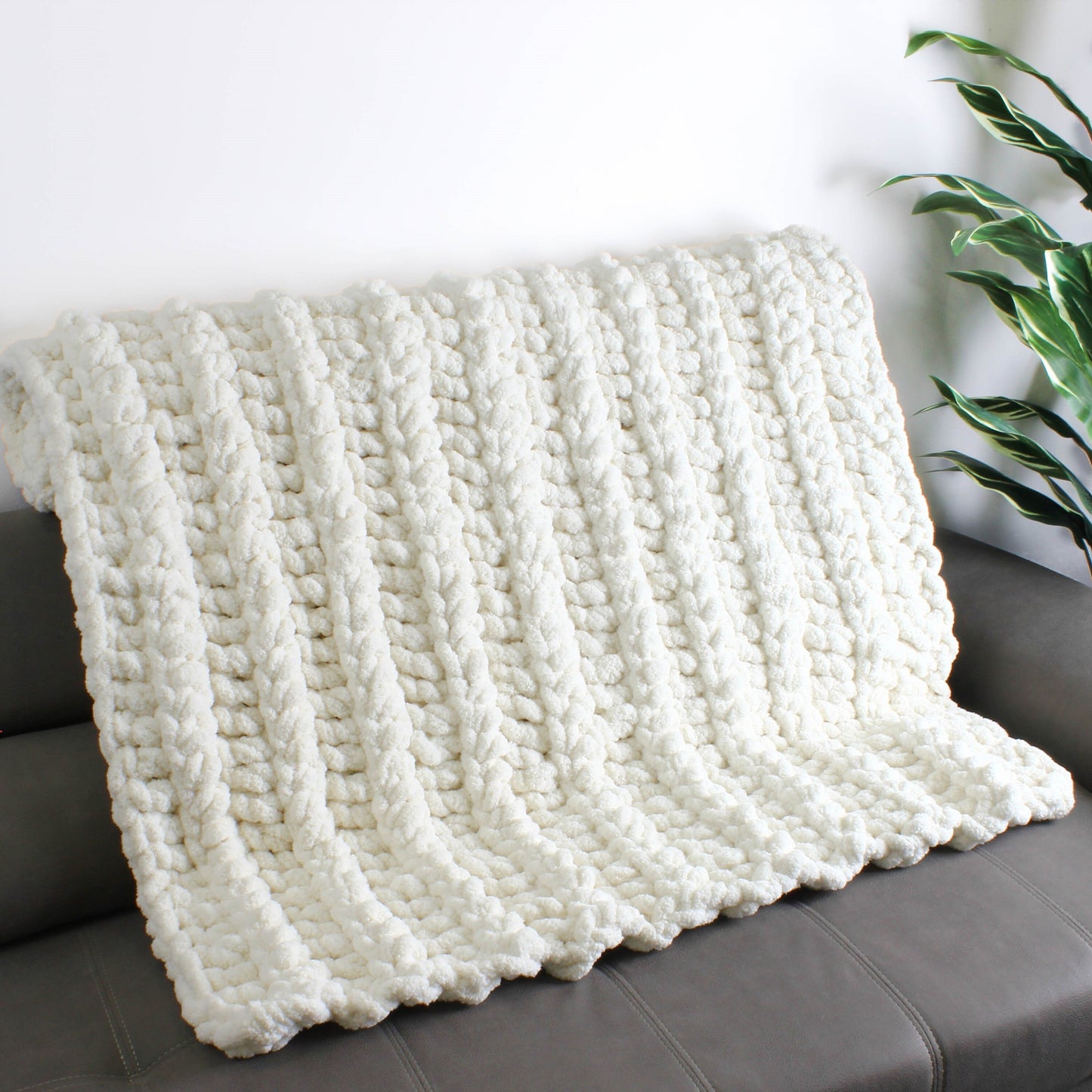 May include: A white knitted throw blanket with a chunky cable knit pattern. The blanket is draped over a gray couch.