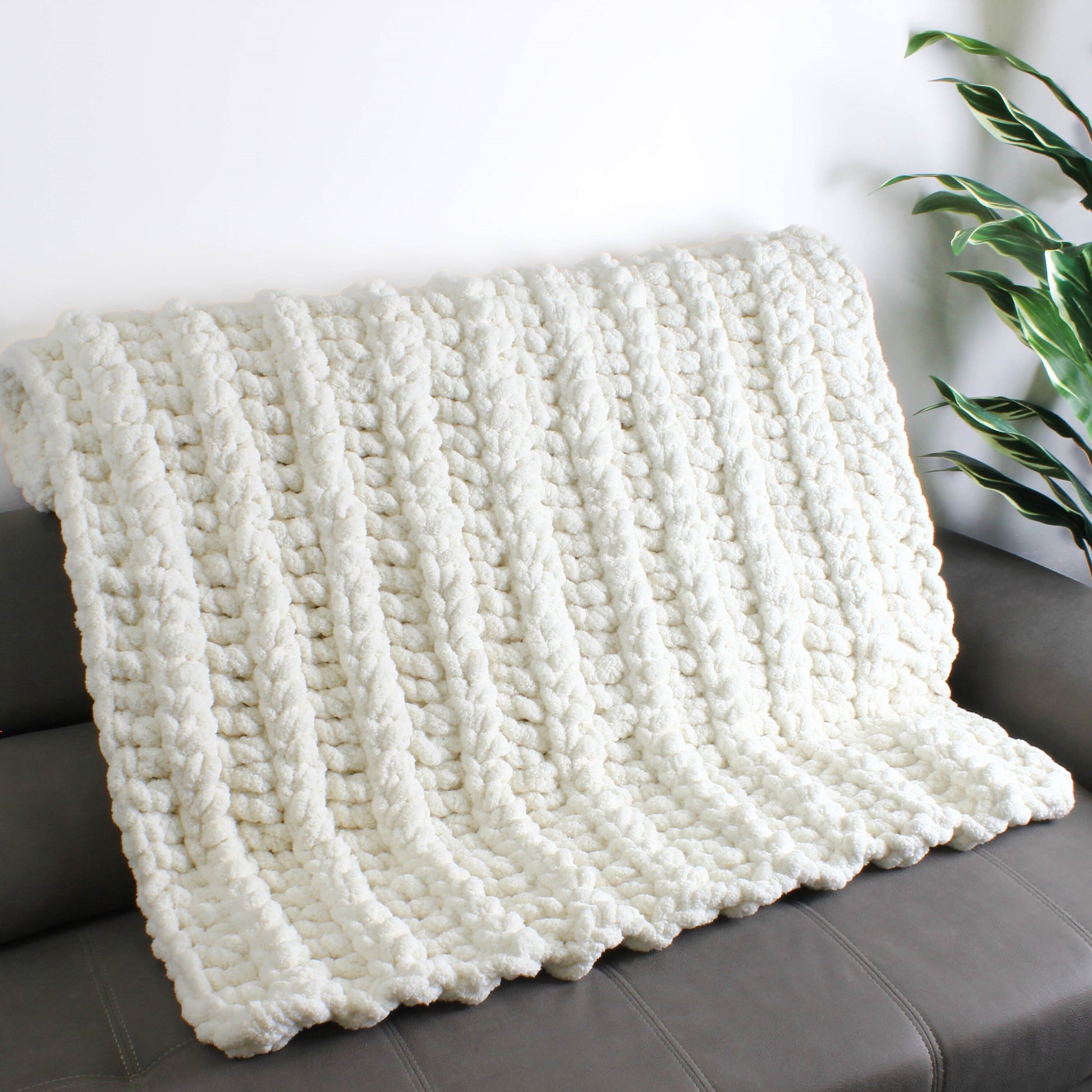 May include: A white knitted throw blanket with a chunky cable knit pattern. The blanket is draped over a gray couch.