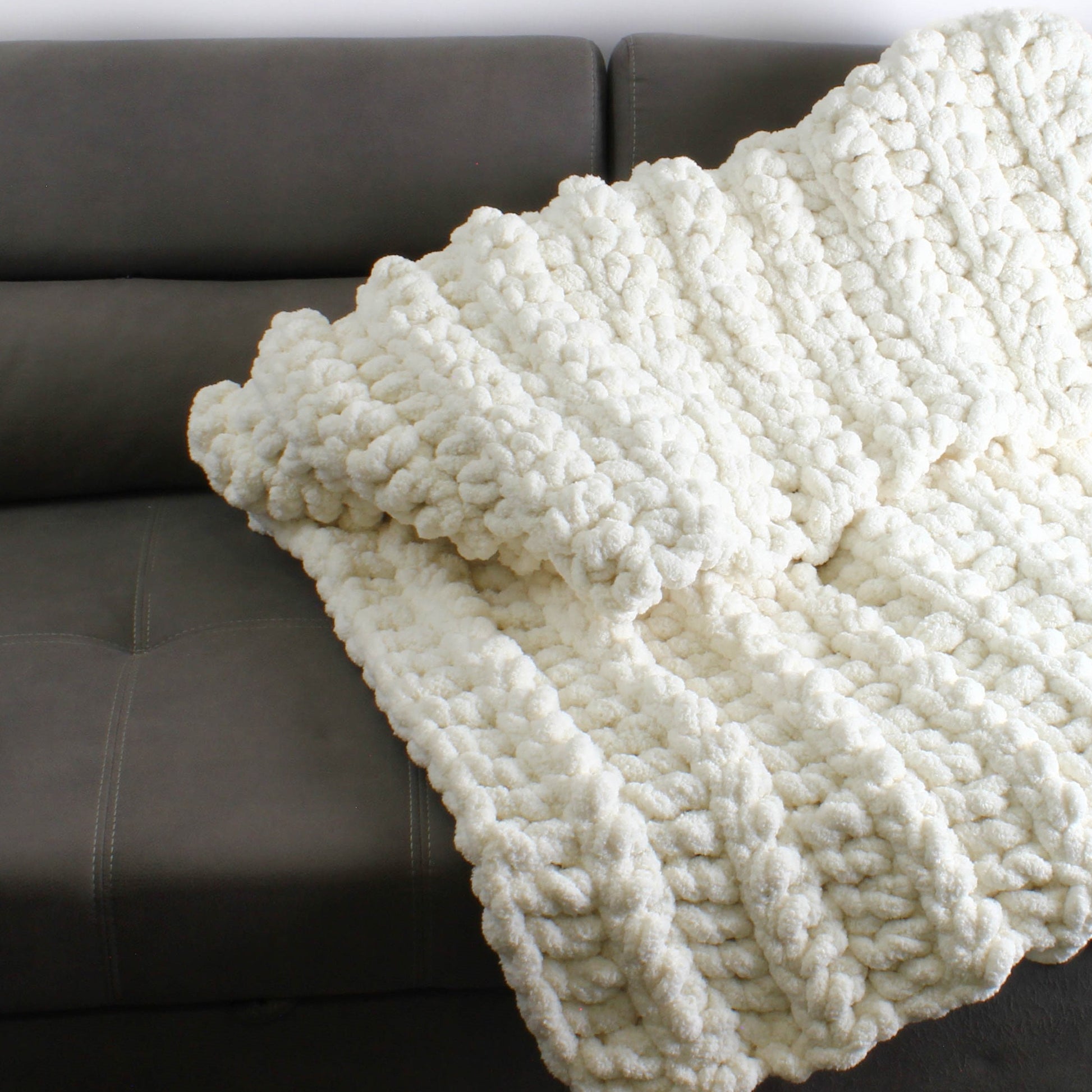 May include: A white chunky knit throw blanket draped over a gray couch. The blanket is made with a thick yarn and has a soft, fluffy texture.