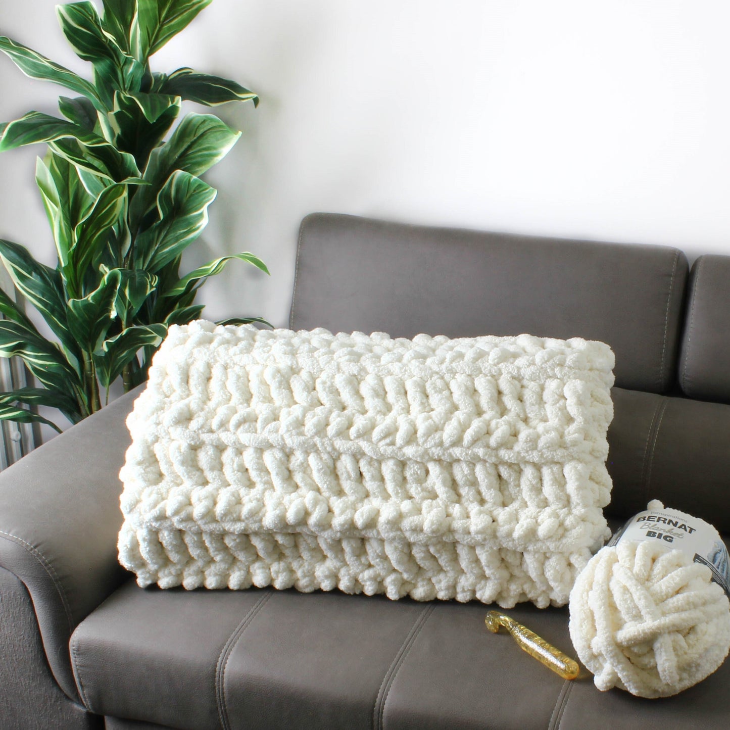 May include: A white chunky knit throw blanket on a gray couch with a ball of white yarn and a crochet hook. The yarn label reads "Bernat Big".