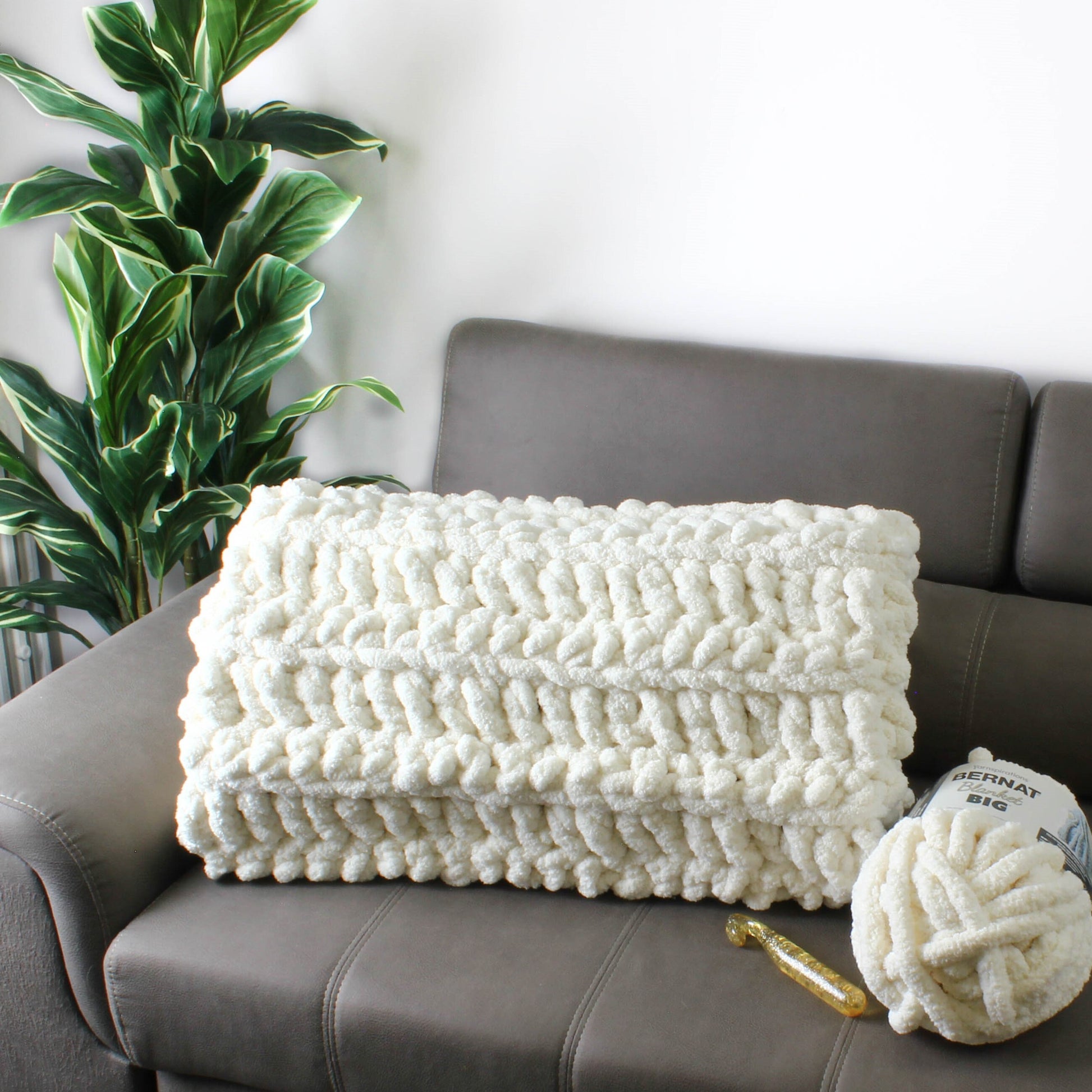 May include: A white chunky knit throw blanket on a gray couch with a ball of white yarn and a crochet hook. The yarn label reads "Bernat Big".