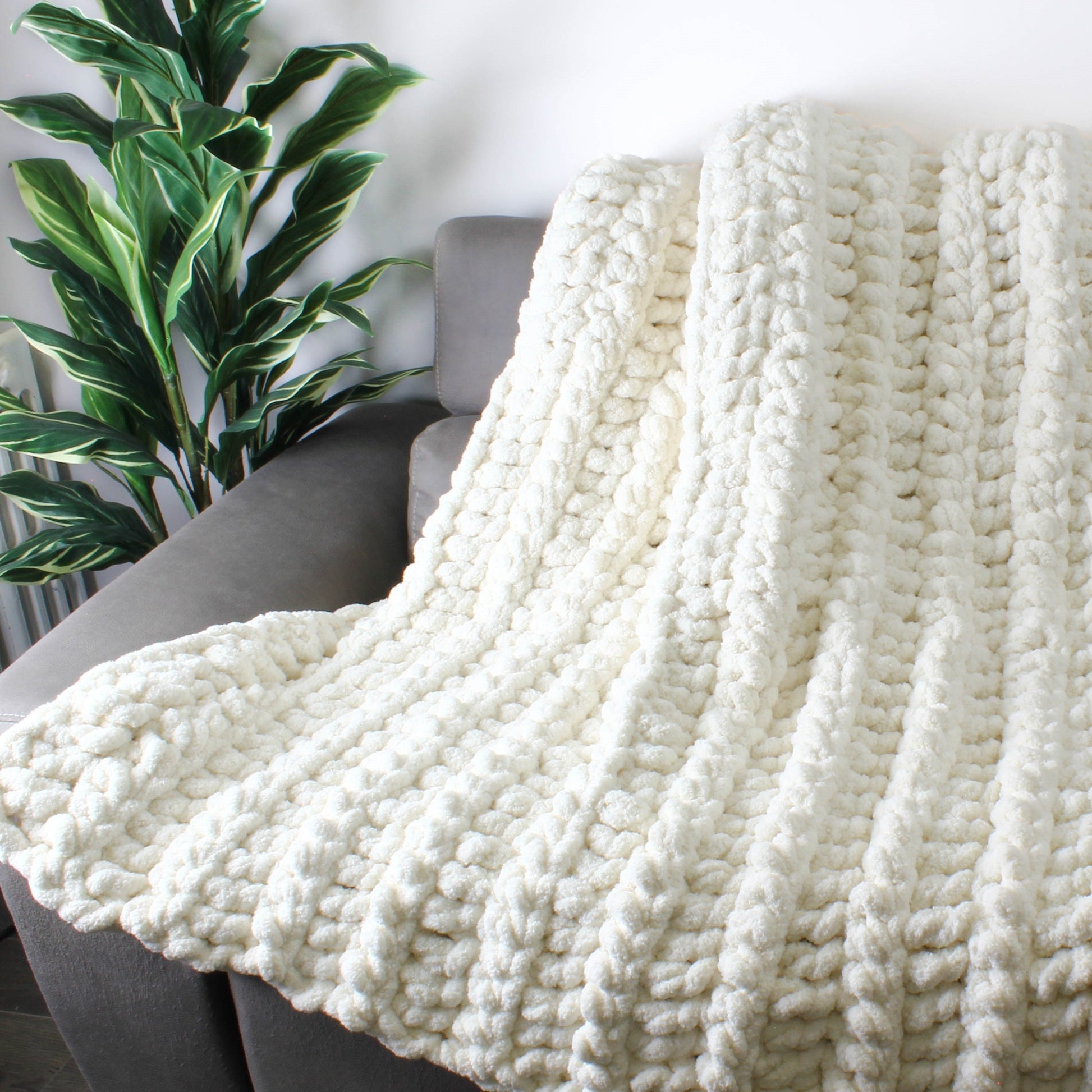May include: A white chunky knit throw blanket draped over a gray couch. The blanket is made with thick yarn and has a soft, fluffy texture.