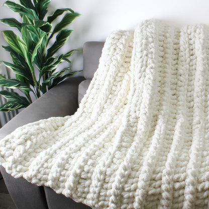 May include: A white chunky knit throw blanket draped over a gray couch. The blanket is made with thick yarn and has a soft, fluffy texture.