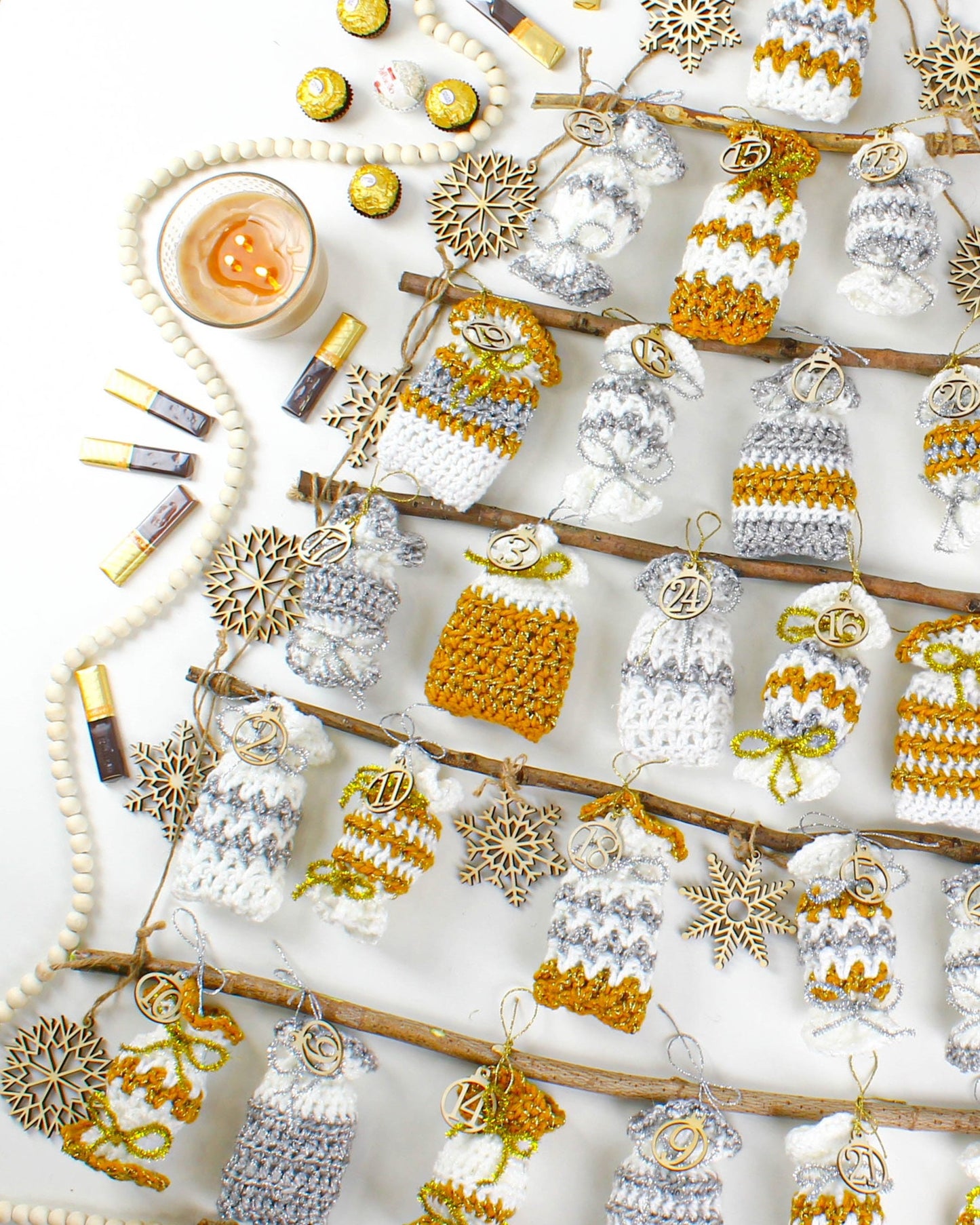 May include: A white background with a wooden branch decorated with crocheted advent calendar pouches. The pouches are in gold and silver with numbers 1 through 24. The pouches are hanging from the branch with twine. There are wooden snowflakes and beads scattered around the branch.