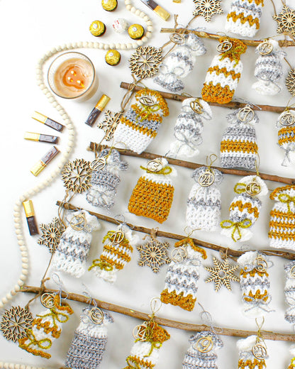 May include: A white background with a wooden branch decorated with crocheted advent calendar pouches. The pouches are in gold and silver with numbers 1 through 24. The pouches are hanging from the branch with twine. There are wooden snowflakes and beads scattered around the branch.