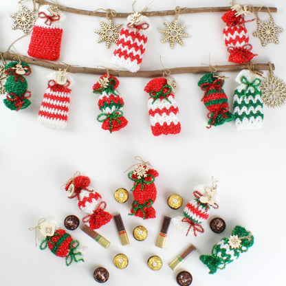 May include: A collection of crocheted candy cane ornaments in red, white, and green, hanging from a wooden branch. The ornaments are decorated with small wooden snowflakes and are surrounded by small chocolate candies.
