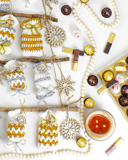 May include: A white background with a Christmas advent calendar made of crocheted pouches with gold and silver accents. The pouches are hung on branches and numbered 16, 15, 22, 21, and 4. There are also wooden snowflakes, a string of wooden beads, and chocolate candies.
