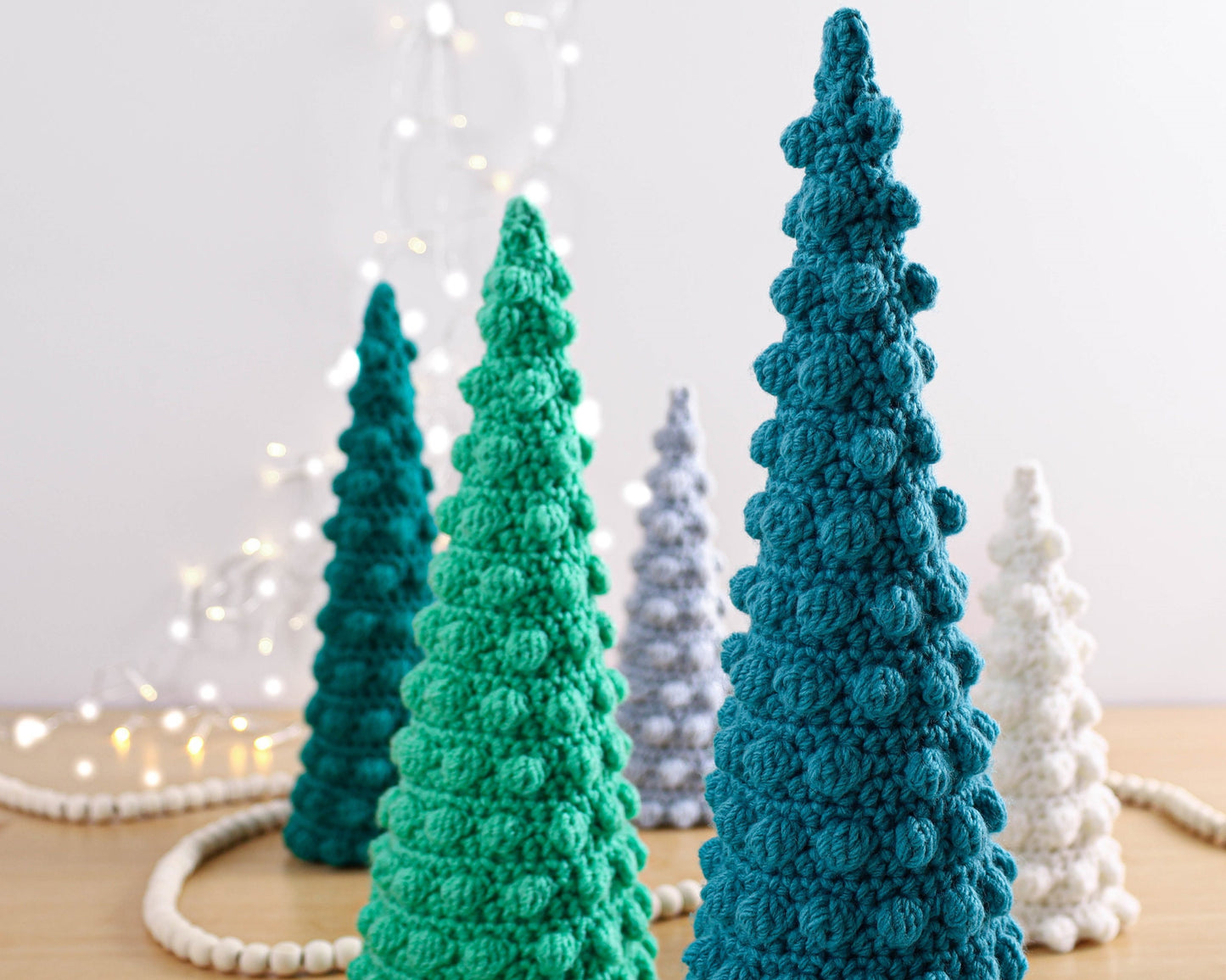 May include: Five crocheted Christmas trees in different colors and sizes. The trees are made with a textured stitch that gives them a bumpy appearance. The trees are arranged in a row on a white surface.