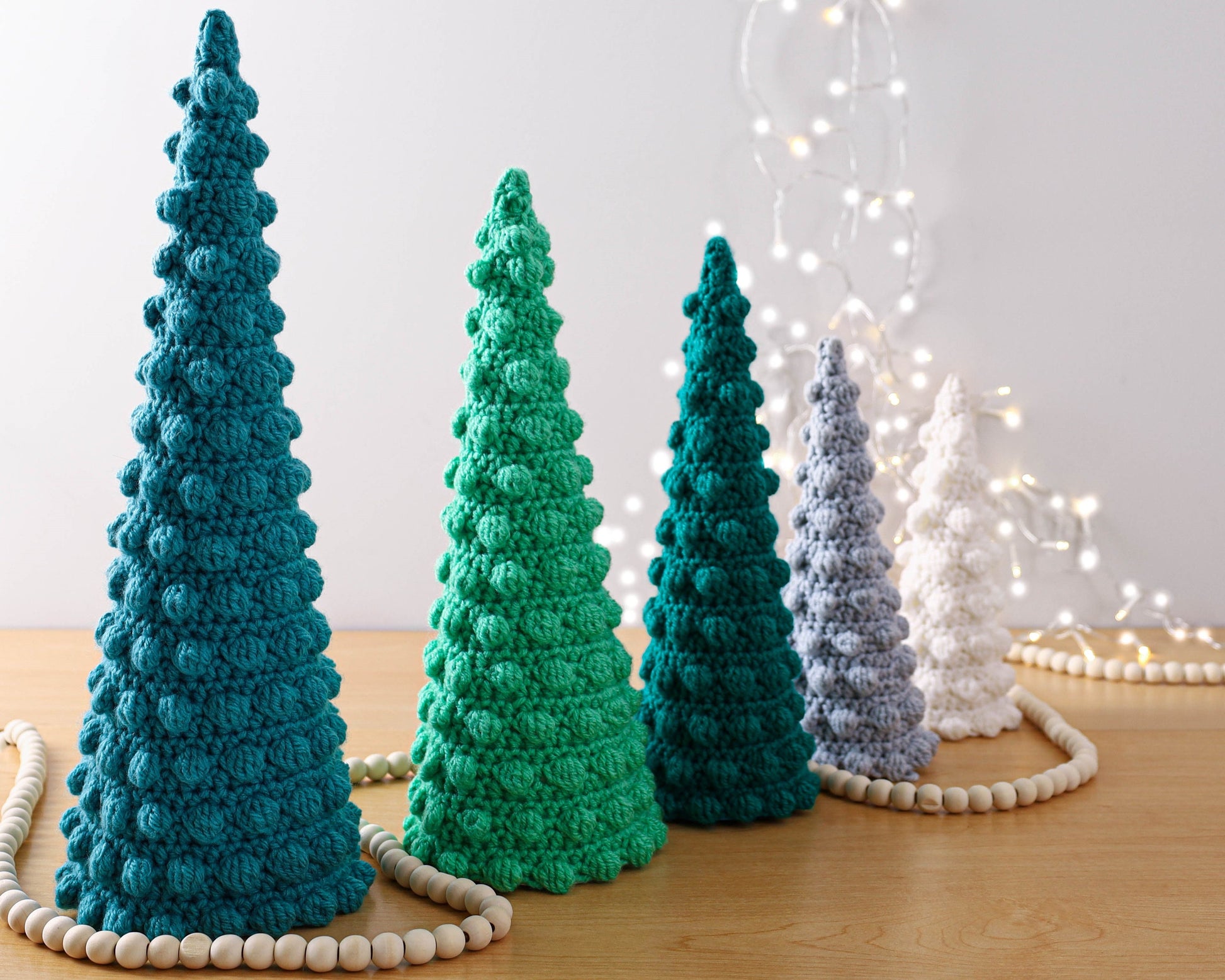 May include: Four crocheted Christmas trees in different sizes and colors: teal, green, gray, and white. The trees are arranged in a row on a wooden surface with white beads scattered around them.