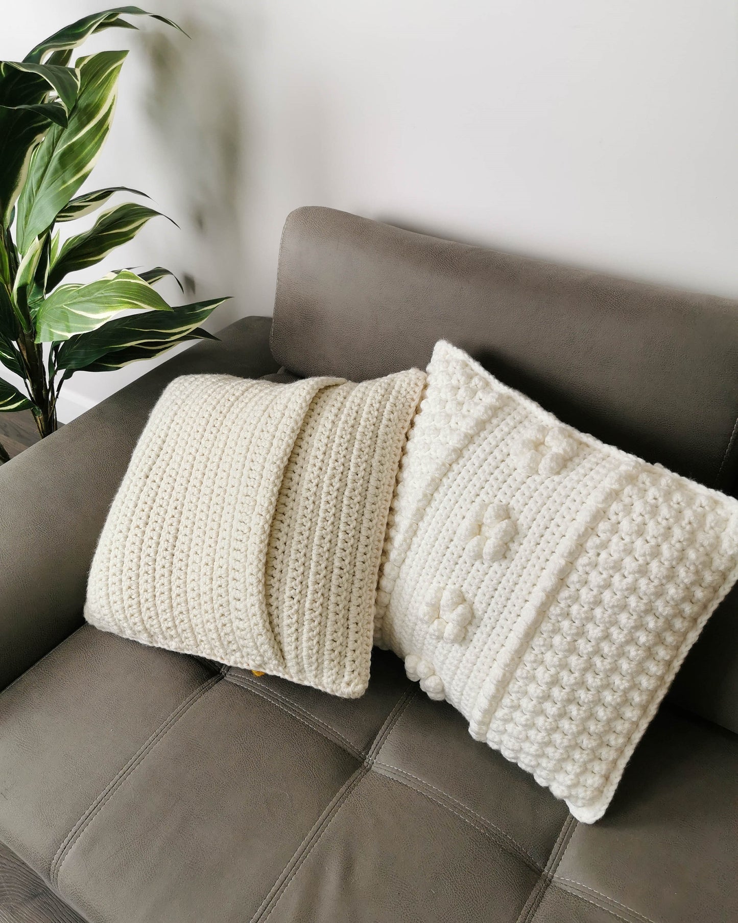 May include: Two white crocheted throw pillows on a brown couch. One pillow has a textured surface and the other has a raised bobble pattern.