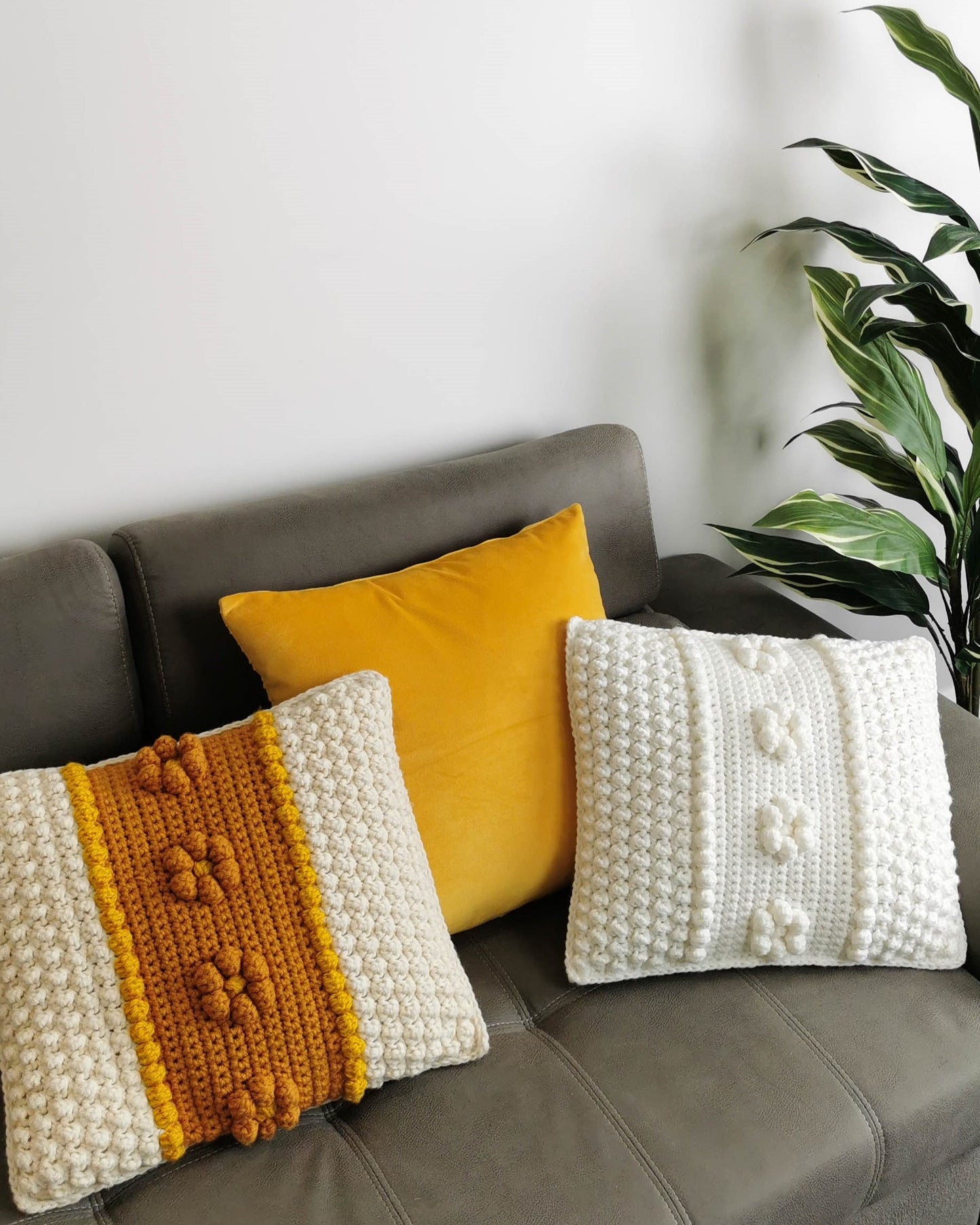 May include: Three decorative throw pillows on a gray couch. One pillow is a solid yellow velvet, and the other two are crocheted with white and mustard yellow yarn.