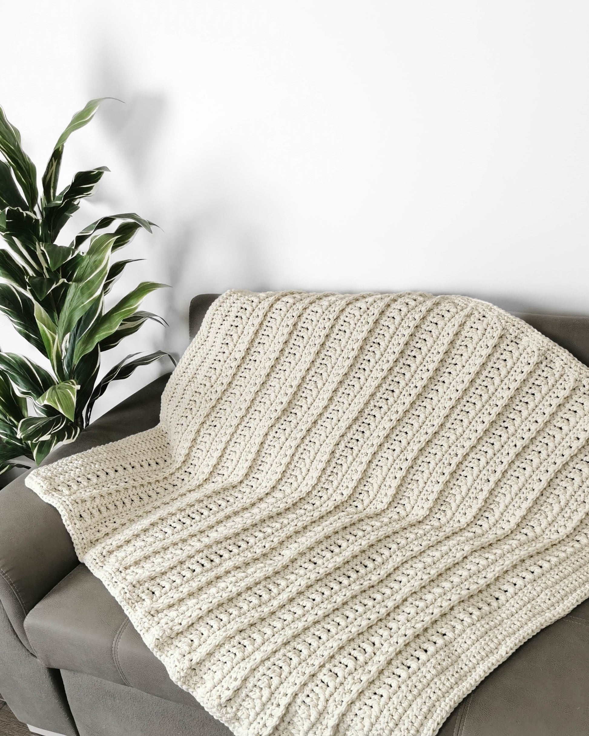 May include: A cream-colored crocheted throw blanket with a ribbed texture is draped over a gray couch.