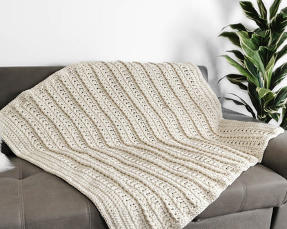May include: A cream-colored crocheted throw blanket draped over a gray couch. The blanket has a textured, ribbed pattern.