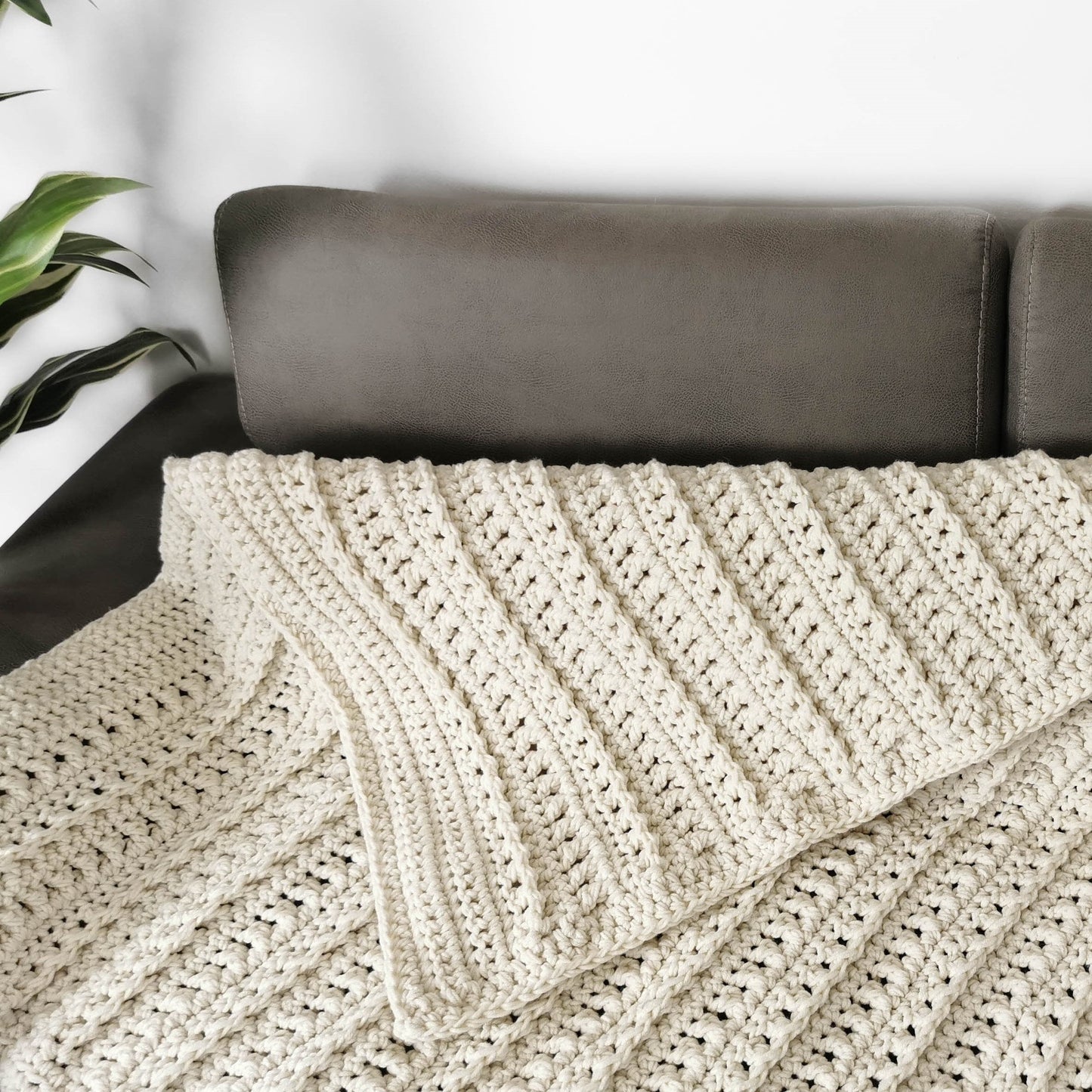 May include: A cream-colored crocheted throw blanket with a textured pattern, folded on a brown couch.