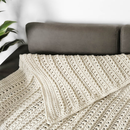 May include: A cream-colored crocheted throw blanket with a textured pattern, folded on a brown couch.